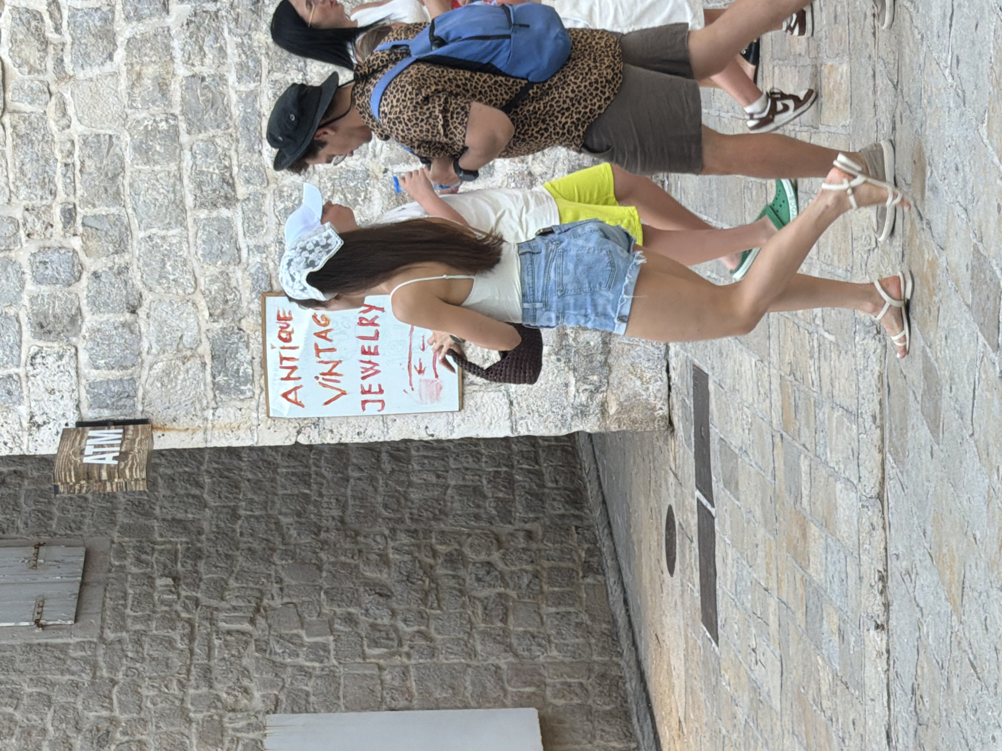 The image depicts a street scene in Budva, Montenegro, likely in the Old Town due to the stone architecture. In the foreground, a young woman in denim shorts and a white tank top walks past a stone wall. She carries a small brown purse and wears white sandals. To her right, a man wearing a leopard-print shirt, dark shorts, a black hat, and a blue backpack walks away from the camera. Behind them, another person in a white shirt and yellow shorts is visible. A sign on the stone wall reads "ANTIQUE VINTAGE JEWELRY" and another sign above it indicates "ATM". The street is paved with stone, and other people are partially visible in the background. The lighting suggests it is daytime.