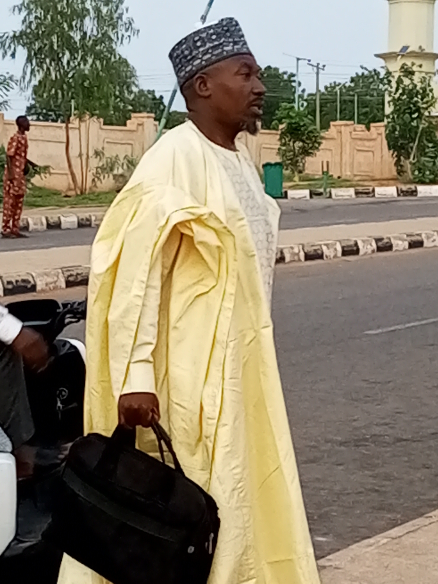 In Gombe, Nigeria, a man in a light yellow traditional robe and a patterned cap stands on a paved road, holding a black duffel bag. He is looking to his right. Behind him, another person in a brown and orange patterned outfit walks away from the camera. The background features a low, beige wall, some trees, and a street lamp. To the right of the main subject, a pale, cylindrical structure with solar panels is visible, possibly part of a building or monument. The road has a white line marking and a raised, patterned curb. A motorcycle is partially visible in the foreground to the left.