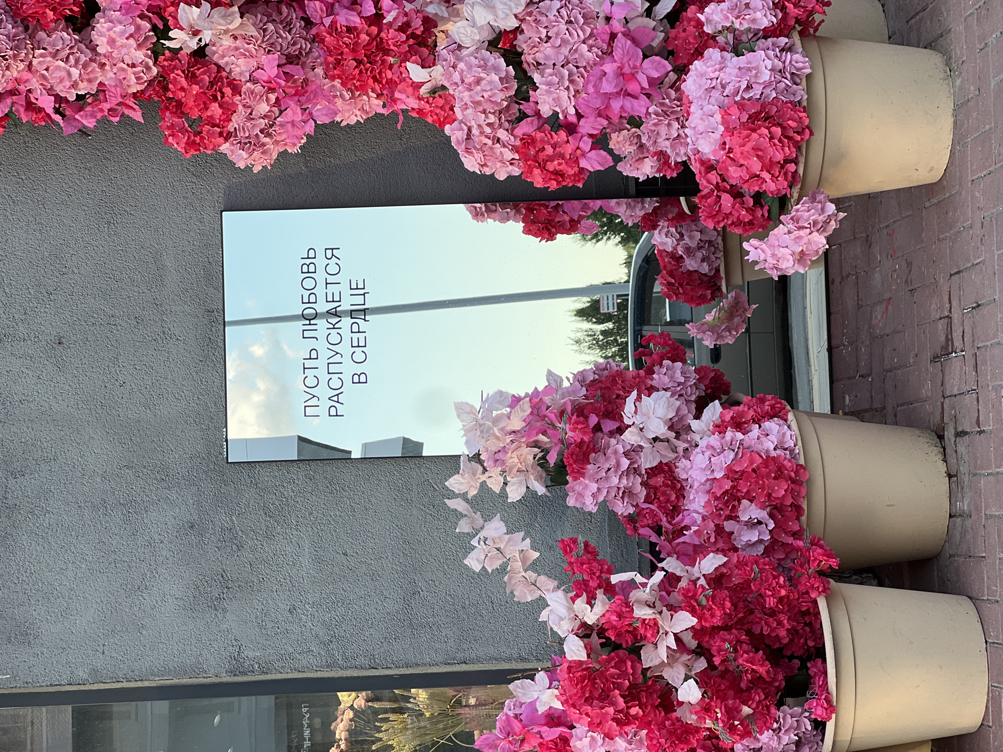 The image shows a decorative display of pink and red artificial flowers against a textured gray wall in Yekaterinburg, Russia. A large rectangular mirror stands upright, reflecting the sky and some trees. The mirror has text written in Cyrillic script: "ПУСТЬ ЛЮБОВЬ РАСПУСКАЕТСЯ В СЕРДЦЕ" which translates to "Let love blossom in the heart." Two large beige planters filled with the same artificial flowers are positioned on the ground in front of the wall. The ground is paved with red bricks. In the reflection of the mirror, a car and a street lamp are visible. The scene appears to be an outdoor setting, possibly a storefront or a public space decorated for an event or as a permanent feature. There are no people visible in the image.
