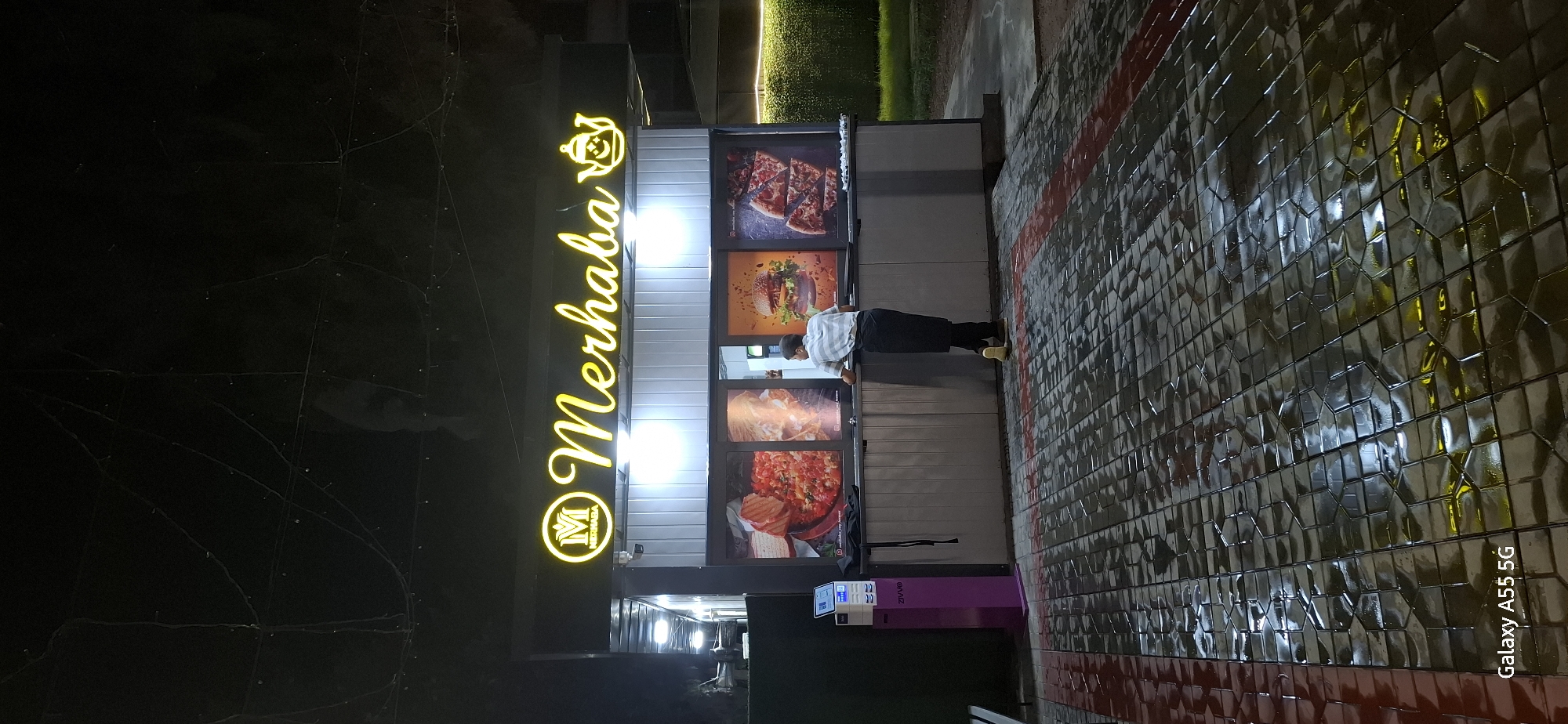 The image shows an outdoor food stand at night in Termez, Uzbekistan. A bright yellow neon sign above the stand reads "Merhaba" with a teapot icon. The stand has a grey corrugated metal facade and displays large posters of food items, including a dish with tomatoes and bread, a burger, and pizza slices. A person, likely an employee, is standing behind the counter, facing away from the camera, looking towards the food displays. They are wearing a light-colored striped shirt and a dark apron. To the left of the counter is a purple payment terminal with a screen. The ground in front of the stand is paved with dark, wet hexagonal tiles, reflecting the lights from the stand and surrounding area. In the background, there are some dimly lit trees and a green hedge. The overall scene suggests a casual dining establishment operating in the evening.