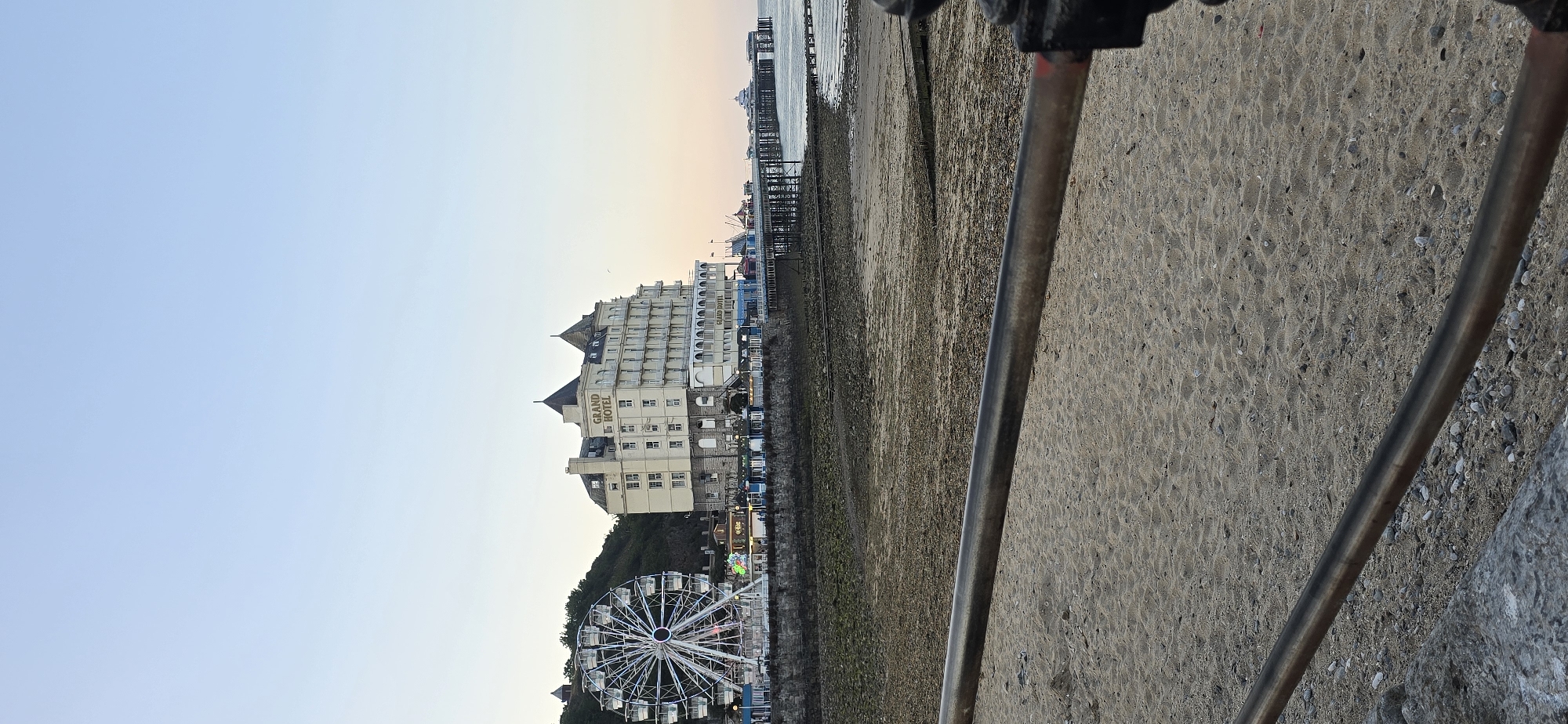 The image captures a view of the Llandudno Pier and the Grand Hotel in Llandudno, United Kingdom, as seen from the beach. In the foreground, a sandy beach is visible, with a metal railing running horizontally across the lower portion of the frame. Beyond the beach, a rocky shoreline leads to the pier, which extends out into the sea. The Grand Hotel, a large, cream-colored building with multiple stories and distinctive turrets, stands prominently next to a Ferris wheel. The Ferris wheel is a large structure with white cabins. Various smaller buildings and amusement stalls are situated along the pier. The sea is calm, with several boats visible in the distance. The sky is clear and a soft blue, indicating it is likely late afternoon or early evening.