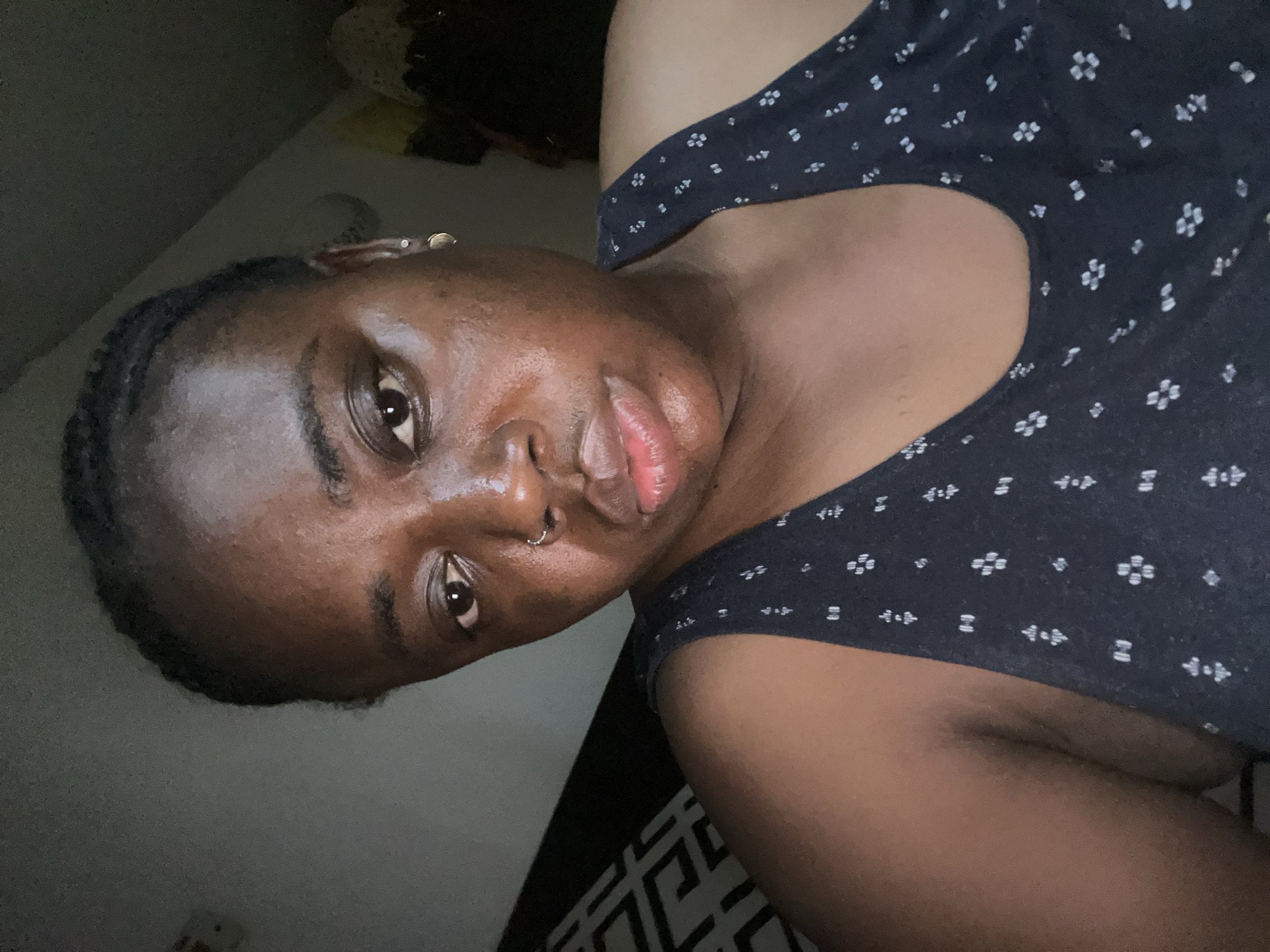 A medium close-up shot shows a dark-skinned woman with braids. She has a nose piercing and a small hoop earring. She is wearing a dark blue tank top with a light-colored floral print. The background is blurred, but it appears to be an indoor setting. The location is not discernible from the image.