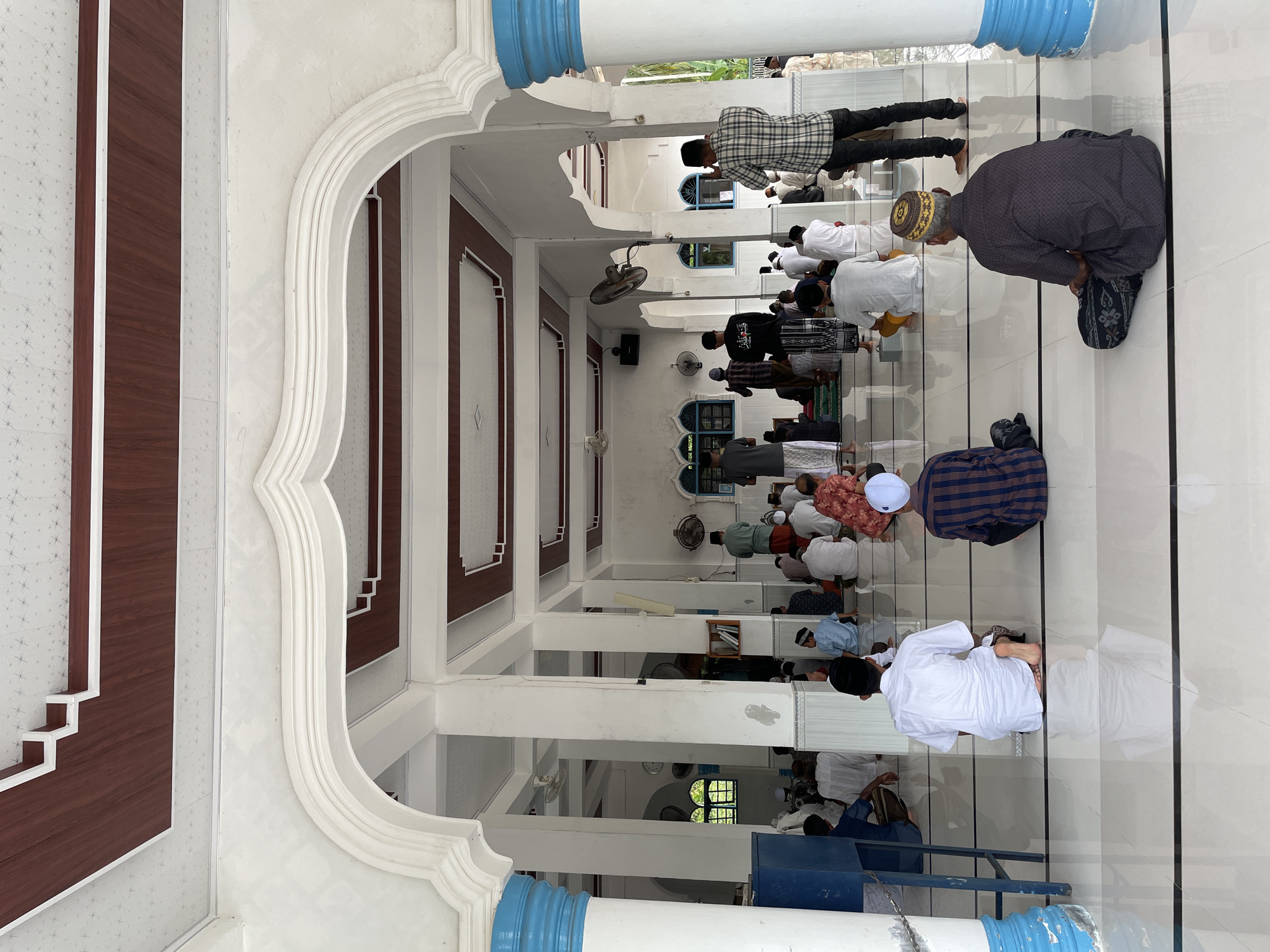 In the city of Trieng Paloh, Indonesia, a group of men are gathered inside a mosque for prayer. The mosque features an arched entryway with white columns, some adorned with blue accents. The ceiling is decorated with wooden panels and white geometric patterns. Inside, men are seated on the floor, facing the front of the mosque, in various stages of prayer. They are dressed in traditional Islamic attire, including sarongs, kufis, and white shirts. Some men are standing, while others are kneeling or bowing. The floor is tiled with white and black lines, reflecting the light from the open space. The environment appears clean and serene, with a few ceiling fans visible and some windows with arched frames in the background.