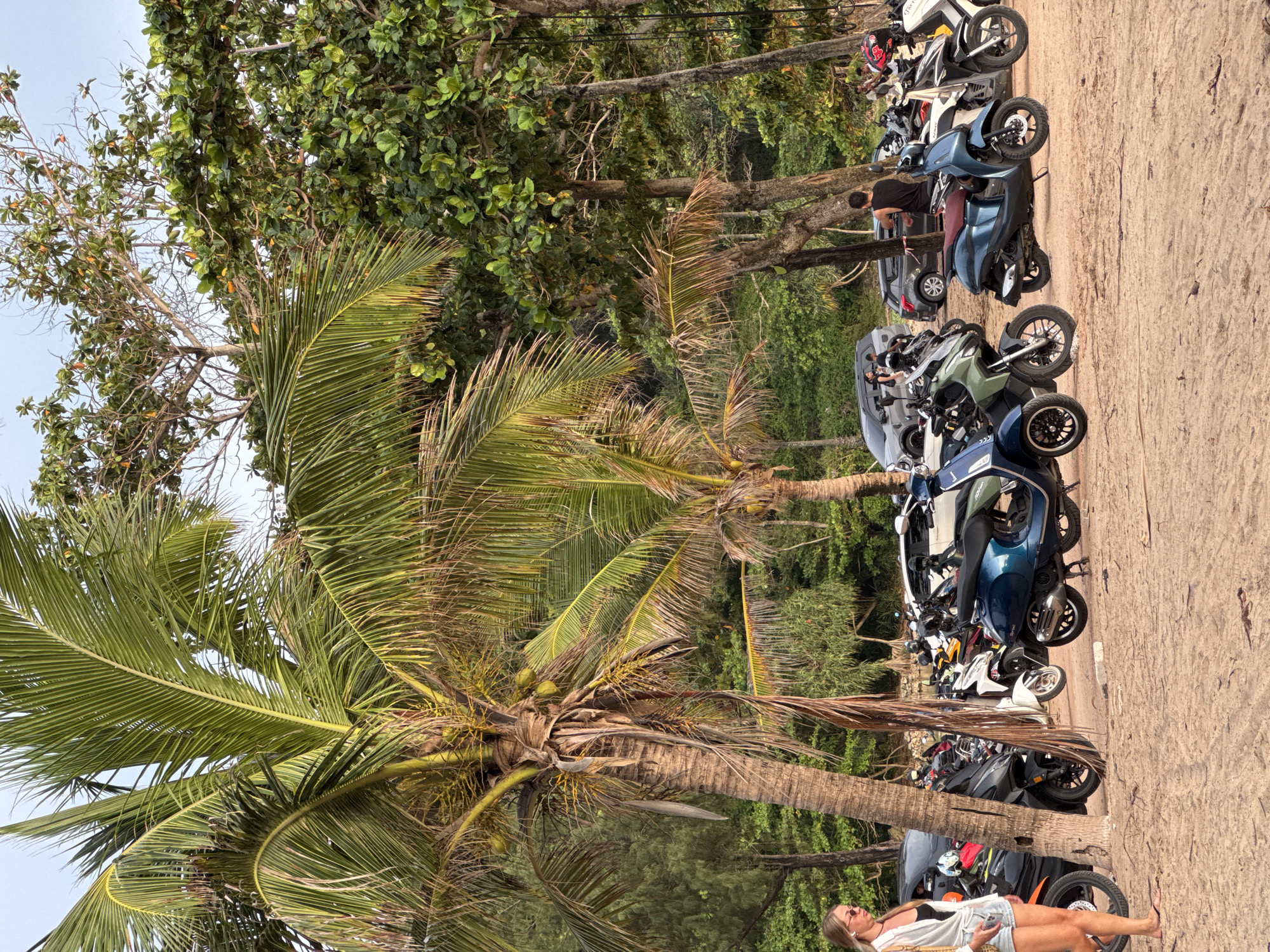 In Rawai, Thailand, a sandy beach area adjacent to a lush green tree line is depicted. Several parked motor scooters and motorcycles are visible, predominantly in shades of blue, green, white, and black. Two individuals are standing, one near the scooters and another further in the background, while a woman in a white shirt and denim shorts walks across the sand in the foreground, carrying a woven bag. A large palm tree with coconuts stands prominently on the left side of the image, with other tropical trees and foliage forming a dense backdrop. The sky is overcast.