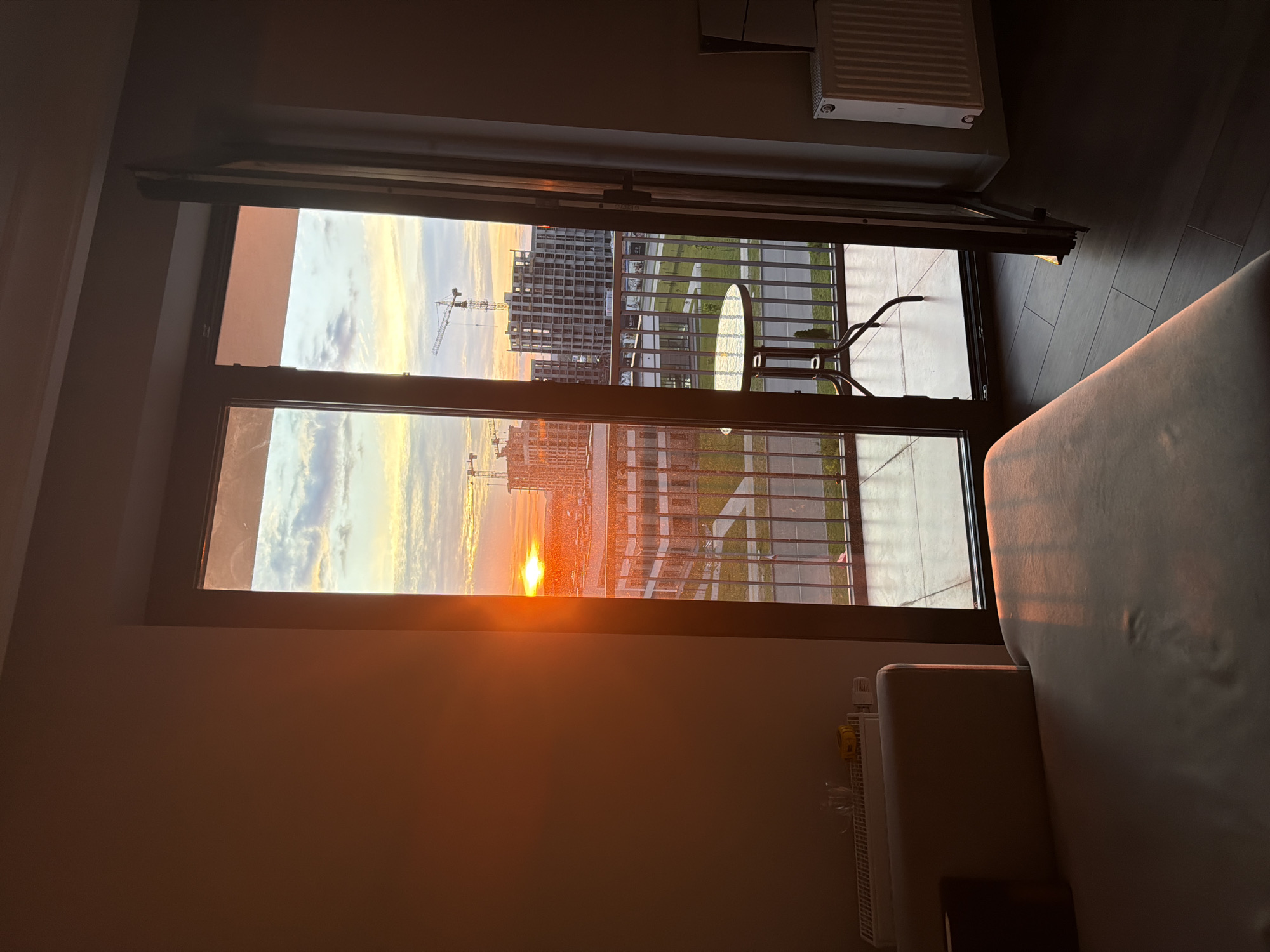 The image displays the interior of a residential room with a direct view of a city landscape at sunset, located in Minsk, Belarus. The setting sun, visible as a bright orange sphere on the left horizon, casts a pronounced golden-orange light across the scene, illuminating surfaces and creating warm highlights.
On the left side of the room, a plain light brown wall extends upwards. A white horizontal panel radiator is positioned at the base of this wall, holding a clear glass pitcher and a small yellow rectangular object. A light-colored sofa, likely beige or light grey, occupies the lower-left foreground, its visible backrest and seat catching the direct sunlight. The floor throughout the room is covered with dark, rectangular wood-effect tiles or laminate.
A large, dark-framed glass door system separates the interior from a balcony in the center. The right-hand door is ajar. On the balcony, a round table with a glass top and dark metal legs stands behind a dark metal railing. The right side of the interior features a darker wall and a white vertical panel radiator. A dark curtain or fabric panel hangs near the open balcony door on this side.
The exterior view reveals an urban environment under a sky transitioning from a vivid orange at the horizon to pale blue with scattered white and grey clouds. Numerous multi-story buildings, many in various stages of construction with visible concrete structures, dominate the middle ground. At least three prominent construction cranes are silhouetted against the sky, indicating ongoing development. Below the buildings, a network of roads, pavements, and patches of green landscaping are visible. No people are discernible in the scene.