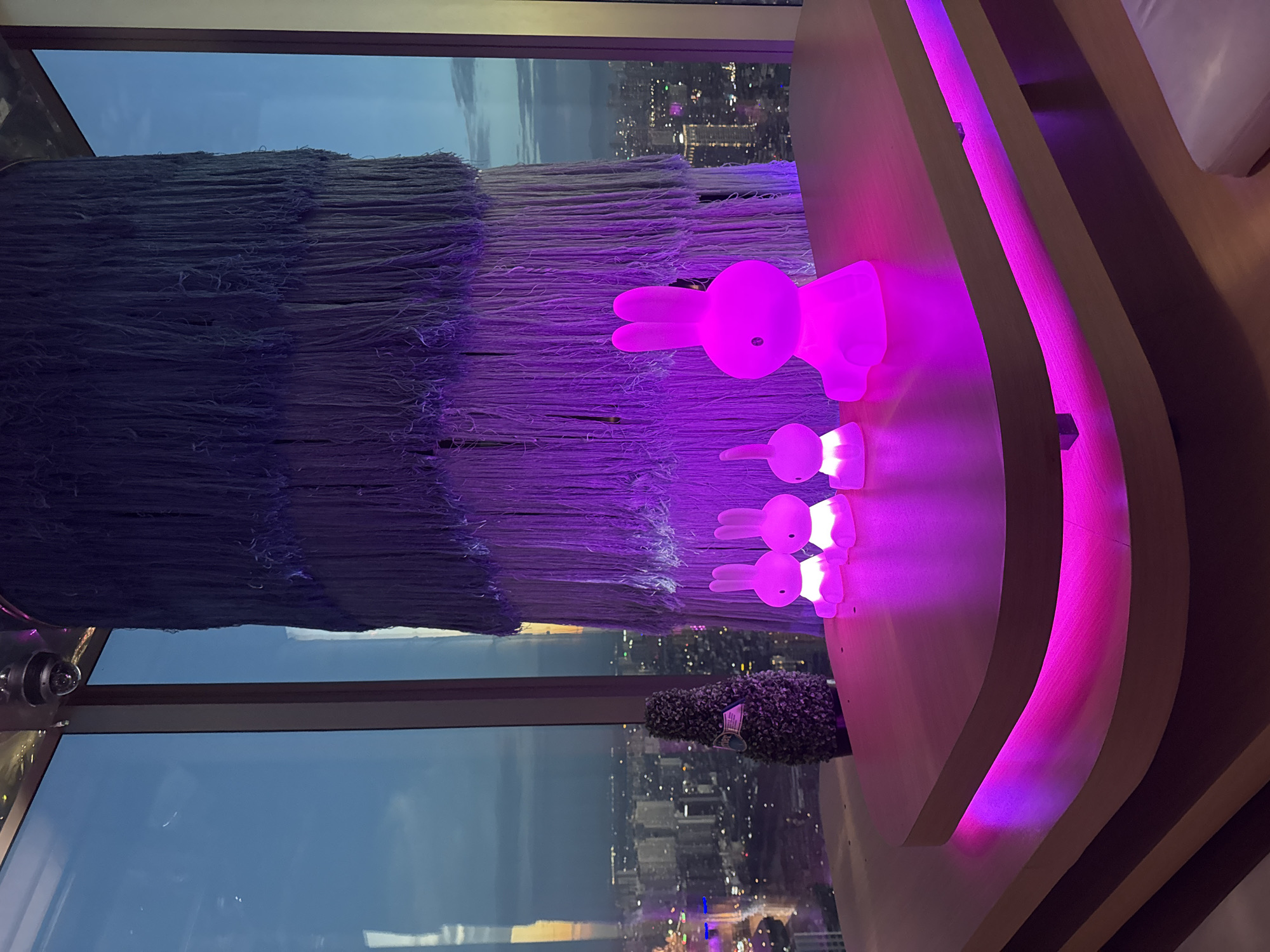 This image captures a contemporary interior design scene likely within a high-rise building in Hanoi, Vietnam. The foreground features a light wood table with a curved edge, illuminated by vibrant pink LED strip lights underneath, creating a soft glow. On the table, several illuminated bunny-shaped lamps are arranged. The largest is a bright pink Miffy lamp on the right, followed by three smaller, similarly shaped lamps to its left, also glowing pink. Behind the lamps, a small, dark green, topiary-like object stands.
The backdrop is dominated by a large window offering a panoramic view of Hanoi at dusk. The city skyline is visible with numerous illuminated buildings and streetlights stretching into the distance. The sky above transitions from a dusky blue to lighter shades near the horizon, with hints of sunset colors.
Hanging from the ceiling in the upper portion of the image is a large, textured decorative element composed of numerous strands of dark blue and purple material, creating a layered, fringe-like effect. A security camera is mounted on the ceiling in the upper left corner. The overall ambiance is modern, stylish, and somewhat whimsical, likely a bar, lounge, or restaurant setting in Hanoi.