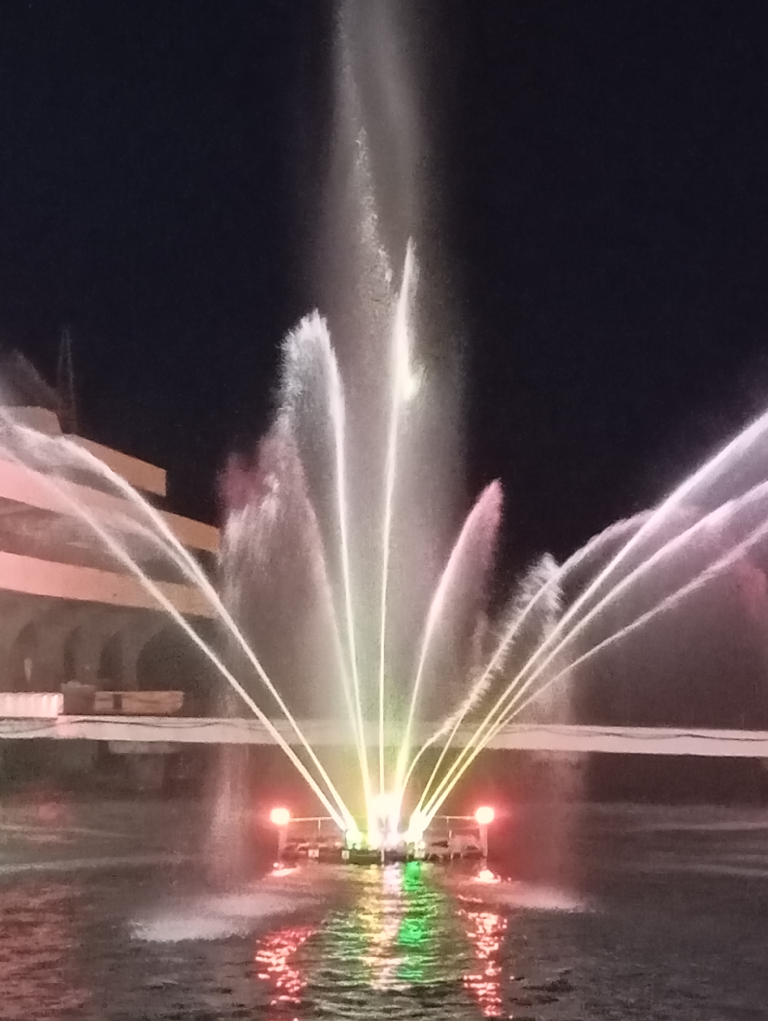 An illuminated fountain display is captured at night. The fountain projects streams of water into the dark sky, with some streams catching colored light in shades of pink, yellow, and green. The water reflects off the surface of the pool, creating a vibrant, shimmering effect. In the background, a modern building with a light-colored facade and arched entrances is visible, suggesting an urban or public space. The overall scene is one of a nighttime attraction, likely in a park or public square, with the fountain serving as the focal point. The location is identified as Sokhumi, Abkhazia.