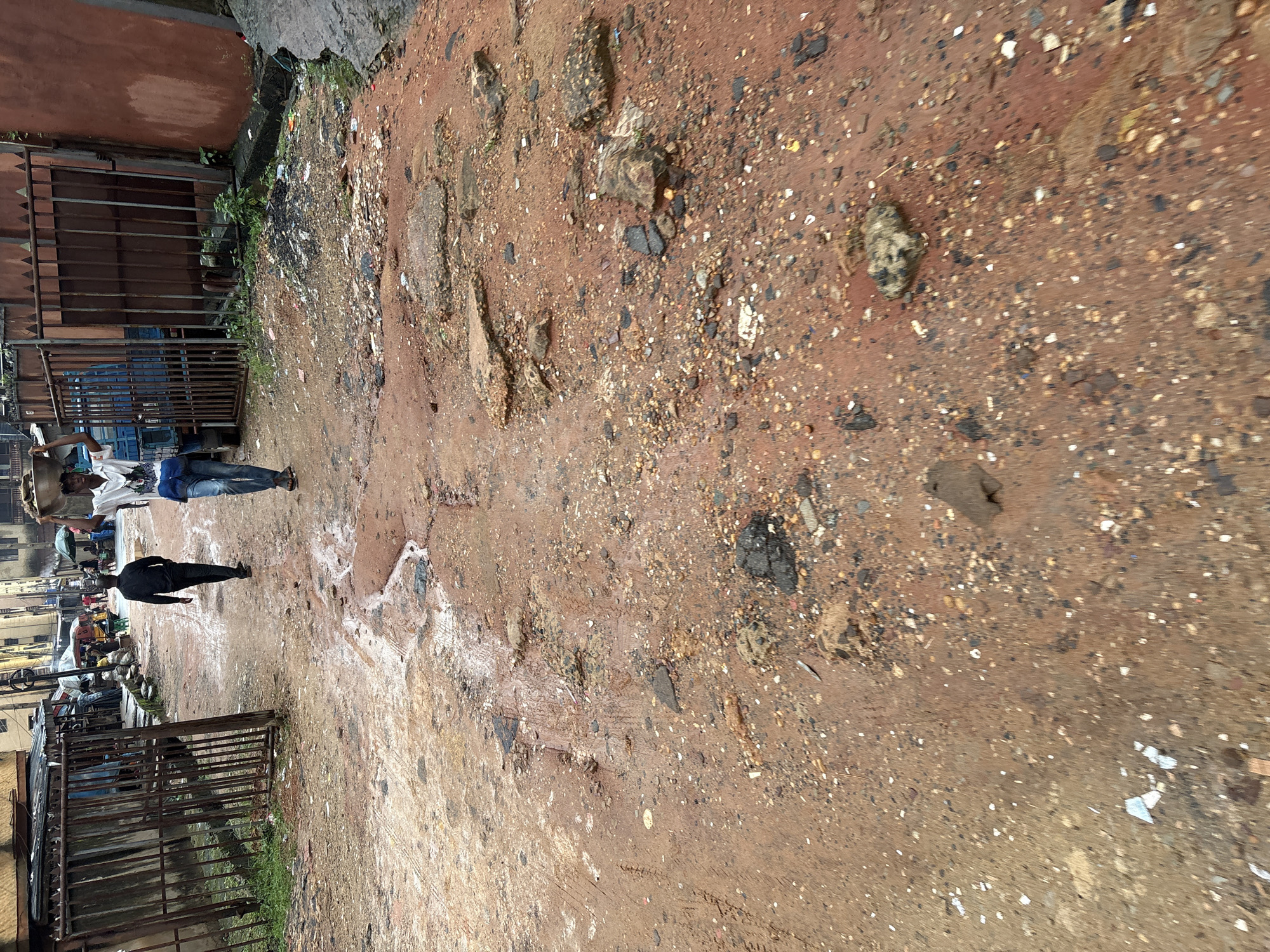 In the city of Onitsha, Nigeria, a two-lane dirt road is heavily deteriorated and riddled with potholes. The road surface is predominantly reddish-brown mud, interspersed with scattered gravel, broken asphalt, and larger stones. Puddles of murky water are present in some of the depressions, indicating recent rainfall or poor drainage.
Two men are walking away from the camera down the road. The man in the foreground is wearing a white short-sleeved shirt with a graphic print on the chest and blue jeans. He is carrying a load on his head. The man behind him is dressed in a black long-sleeved shirt and dark pants.
To the left of the road, a weathered metal gate with vertical bars is partially obscured by overgrown green grass. Beyond the gate, a row of stalls and market structures can be seen, with some people milling about. A blue truck is parked behind the gate.
To the right of the road, a reddish-brown wall runs along the edge. More debris and vegetation are visible near the base of the wall. The overall environment appears to be an urban setting with signs of underdevelopment and neglect.