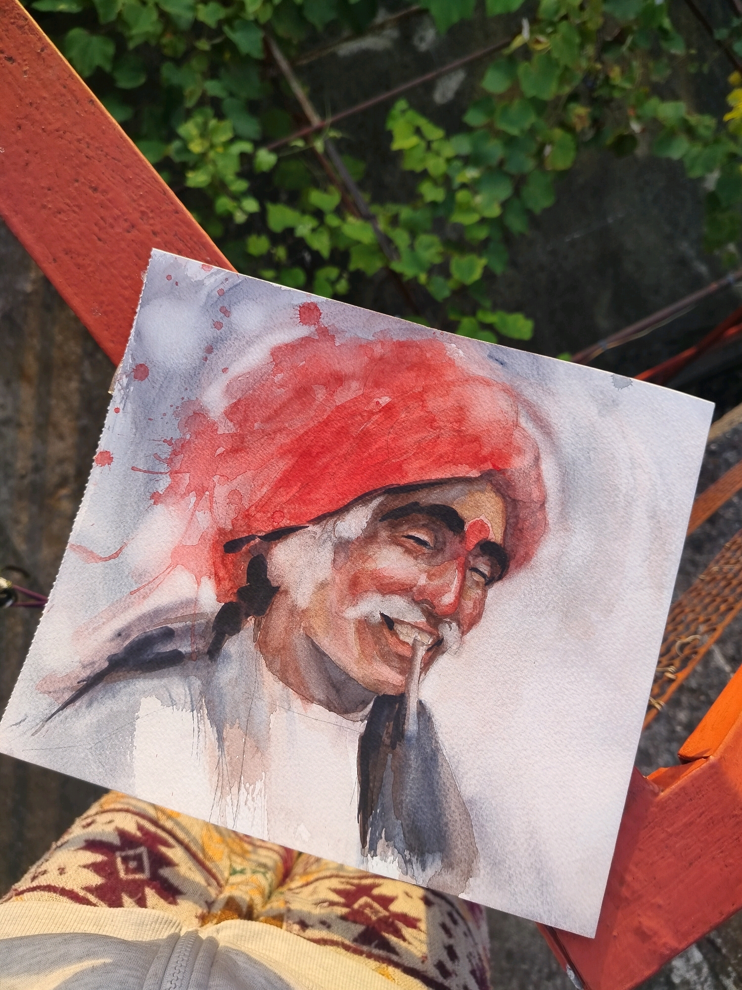 A watercolor painting of an elderly man wearing a red turban is displayed outdoors. The painting is on a square piece of paper, slightly angled, and partially covers the lap of a person wearing a patterned garment with a visible zipper. The man in the painting has his eyes closed and is smiling, with a piece of material visible in his mouth. He has a red bindi on his forehead and a white mustache. The background of the painting is a wash of gray and white with red splatters around the turban. The man's clothing is depicted in shades of gray and brown. The painting is positioned on an orange metal railing, with green foliage and a concrete wall visible in the background. The lighting suggests it is daytime, with soft shadows indicating a slightly overcast or late afternoon sun. The overall scene suggests a casual outdoor setting where the painting is being presented or photographed.