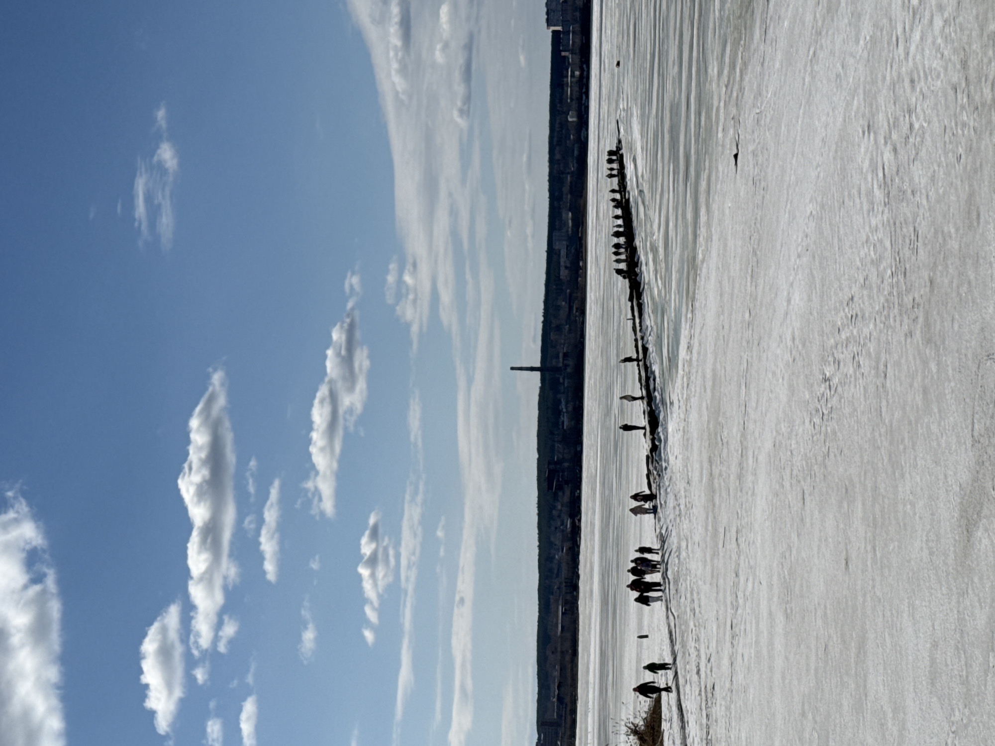 The image depicts a frozen body of water, possibly a lake or river, adjacent to a city skyline. A line of structures or possibly a pier extends into the water from the shoreline. A large number of people are visible on the frozen surface, primarily near the edge, with some individuals more distant.
The city skyline includes buildings of varying heights, and what appears to be a tall smokestack is a prominent feature. The sky is a clear blue with scattered white clouds. The light suggests it is daytime.
Based on the general appearance and architectural style of the buildings, along with the distinctive smokestack, the location is likely Izhevsk, Russia.