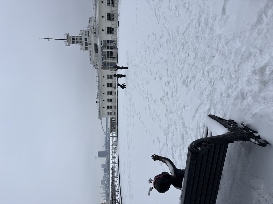 The image displays a snow-covered winter scene at the Kyiv River Port, located on Poshtova Ploshcha in Kyiv, Ukraine, under an overcast grey sky.
In the immediate foreground, the back of a dark bronze child statue is visible, seated on a dark, snow-dusted park bench. The statue wears a red headband adorned with two small white deer-like figures, and its right arm is raised with an open hand. The ground around the bench is covered in fresh snow with visible footprints.
The midground features a wide, flat expanse completely covered in undisturbed snow, also marked by numerous footprints. Three additional dark bronze child statues are positioned within this area: one statue is in a kneeling or crouching posture, while the other two stand upright, both with arms raised, one higher than the other.
The background is dominated by the Kyiv River Port building, a distinctive white, multi-story structure with a ship-like architectural design and a prominent central tower resembling a bridge mast. The text "American University Kyiv" is visible on the building's facade. Beyond the port building, a distant city skyline is discernible across what appears to be the Dnieper River, showing various buildings, construction cranes, and a bridge under the same grey sky.