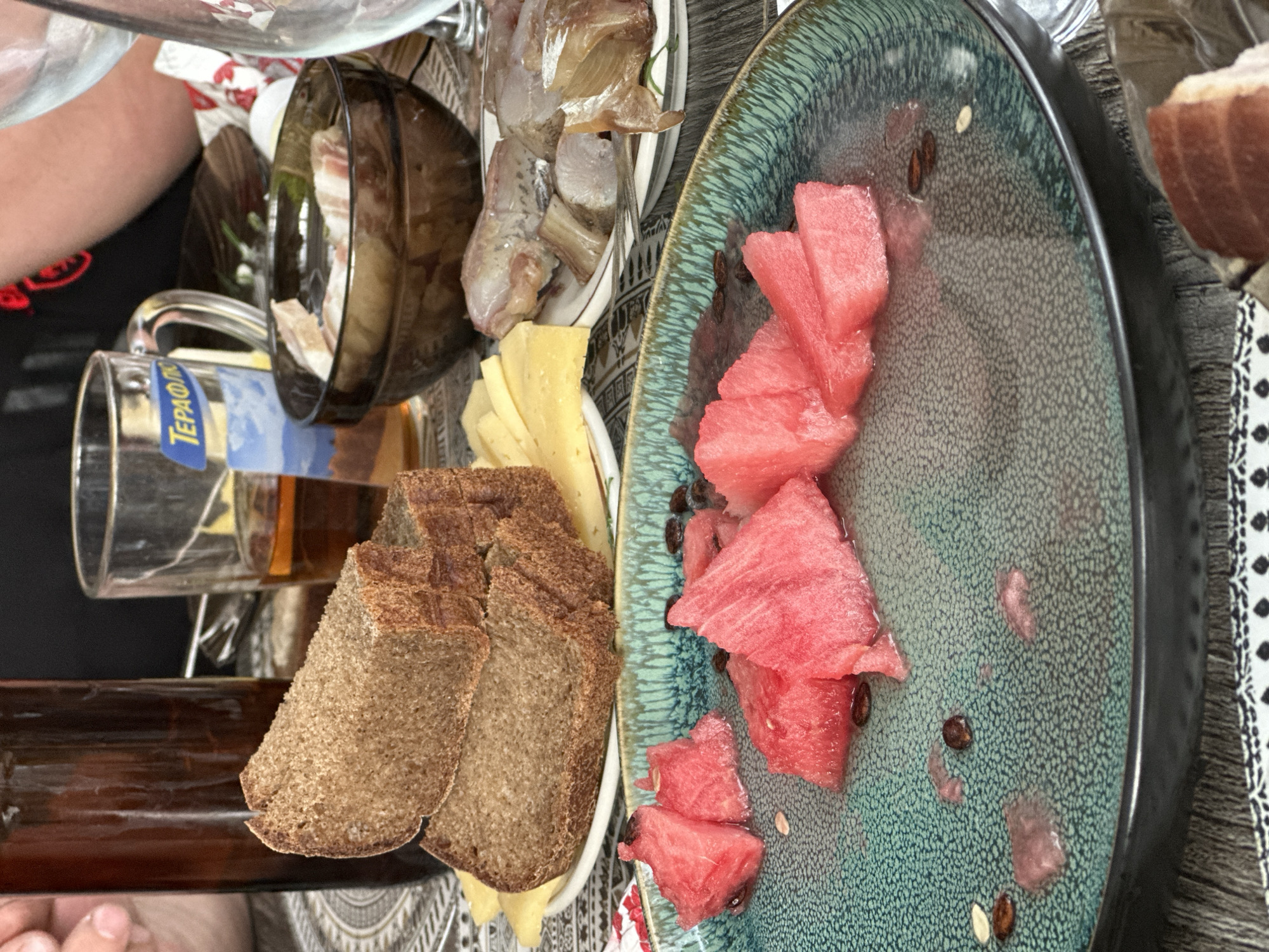 This close-up, eye-level shot captures a table setting with a meal in progress. The foreground features a turquoise-colored plate with several triangular slices of watermelon, some with visible seeds and juice. To the left, a hand is partially visible, reaching towards a dark brown glass bottle, possibly containing a beverage. Behind the watermelon plate, a white plate holds slices of yellow cheese and two thick slices of dark bread. Further back, a dark brown bowl contains pieces of what appears to be cured meat or bacon. A clear glass mug with a blue and white label is also present, containing a light brown liquid, likely beer. In the background, more plates and glasses are visible, suggesting a shared meal. The overall setting appears to be outdoors, on a wooden surface, with a patterned tablecloth underneath. The image evokes a casual dining experience. The location is identified as Baklashi, Russia.
