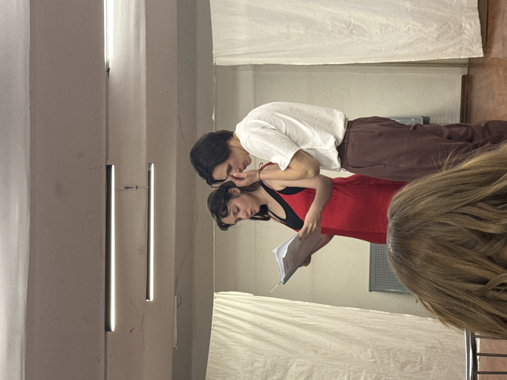 Two people, a man and a woman, are closely positioned in what appears to be an indoor setting. The man is wearing a white shirt and brown pants. He has dark hair and appears to be whispering into his hand, near his face. The woman is wearing a red dress with black accents. She is holding what looks like a script or papers in her hand. The environment has a neutral, somewhat unfinished or rehearsal-like quality, with white curtains or fabric panels in the background. Vertical lighting fixtures are visible on the wall. The image is focused on the interaction between the two individuals, which is not clearly Moscow, Russia based on the objects.