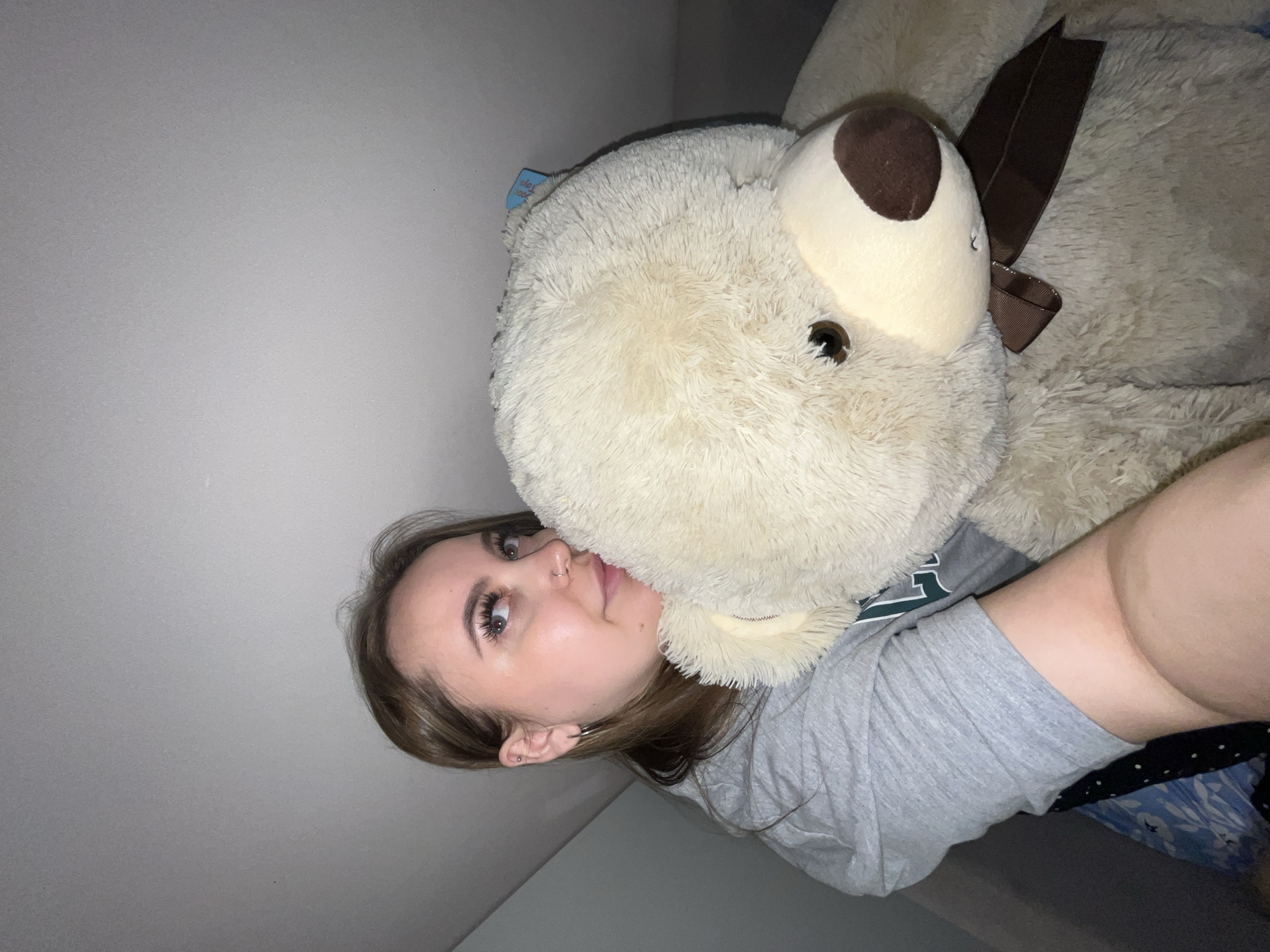 A young woman is holding a large, beige teddy bear. She is wearing a gray t-shirt, and her face is close to the bear's face. The background consists of a white wall and what appears to be a bed with blue patterned sheets. The location cannot be determined from the visual information provided.