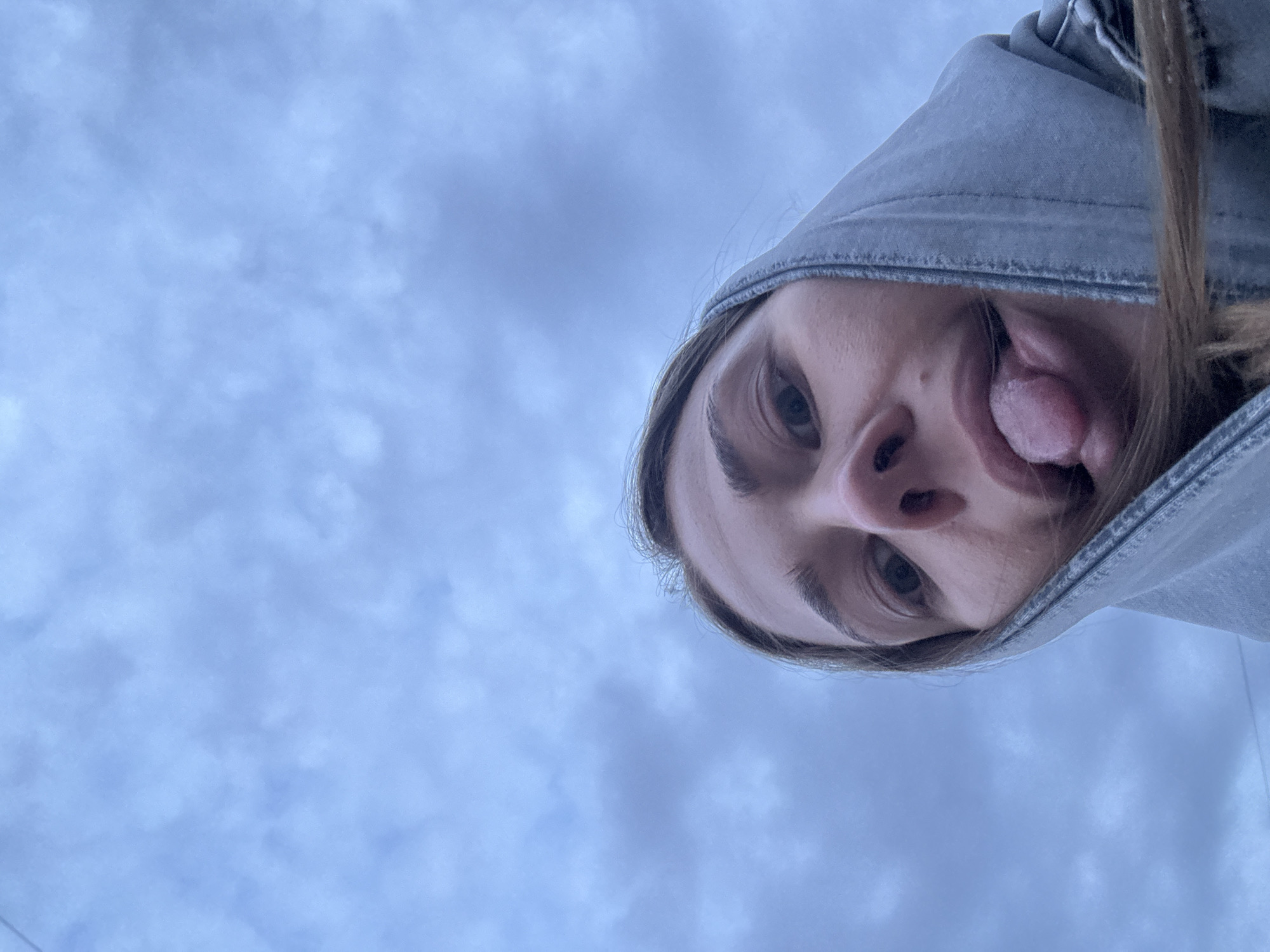 The image presents a rotated, extreme low-angle close-up of a young individual, likely female, looking down towards the camera from an upward perspective. The person's head is tilted diagonally across the upper right portion of the frame, partially obscured by the hood of a greyish-blue garment, possibly a hoodie or jacket. She is captured mid-expression, playfully sticking her tongue out to the right side of her face. Her eyes are visible, along with well-defined eyebrows and parts of her nose and mouth. Light brown hair is visible around her face, emerging from the hood. The background is an overcast sky, characterized by soft, diffused light blue and light grey clouds, indicating a cloudy daytime setting. No harsh shadows are present, suggesting even lighting. The overall mood is candid and lighthearted. No specific location cues are visible within the image itself, but the provided context places the scene in Chernushka, Russia. No text is visible.