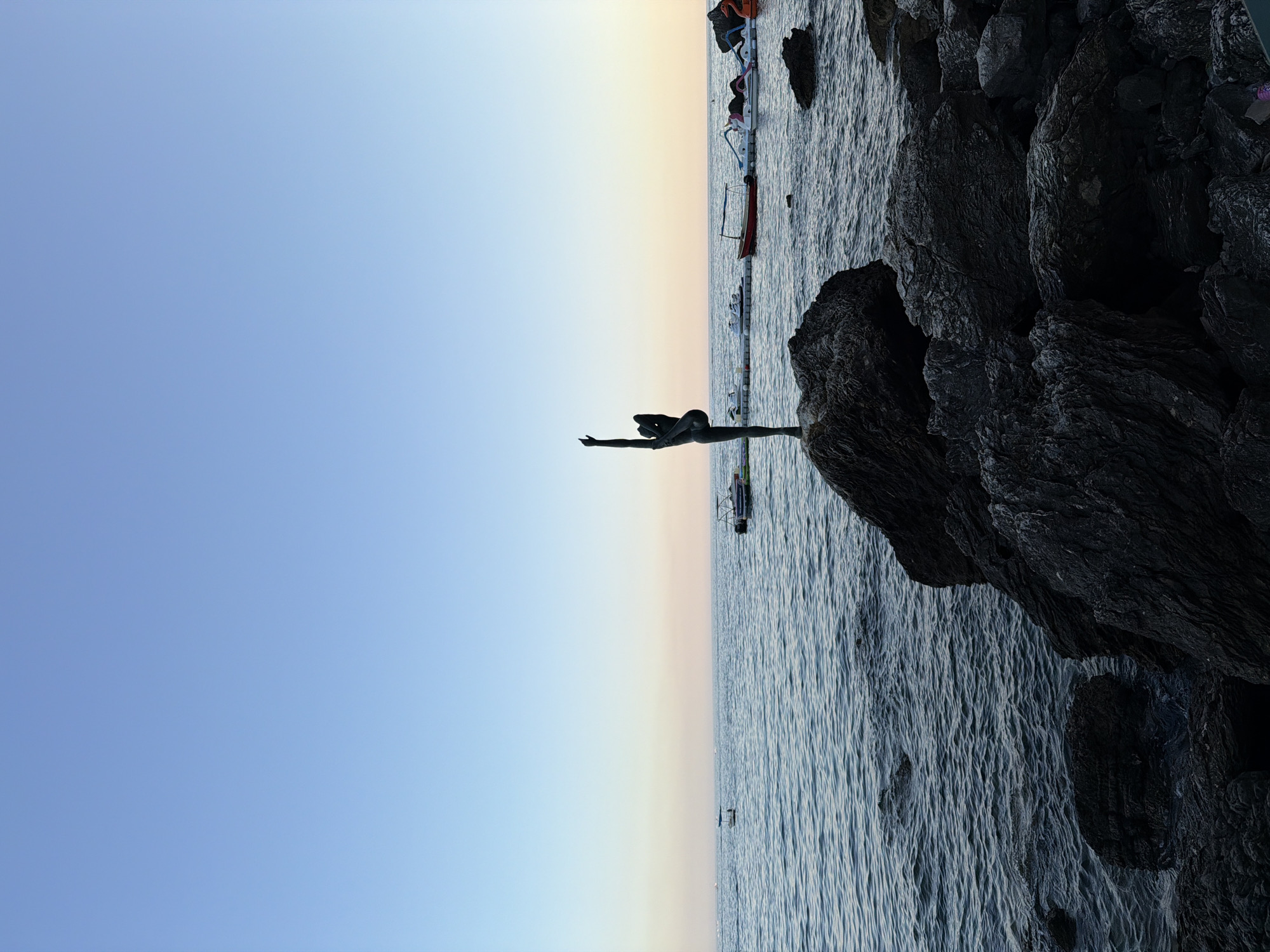 A bronze statue of a ballet dancer in a graceful pose stands atop a large, dark rock formation on the coast of Budva, Montenegro. The statue is silhouetted against a clear blue sky that transitions to a pale pink and orange hue near the horizon, indicating either sunrise or sunset. The sea, with gentle waves, stretches out before the statue. In the background, several boats are moored near a floating dock, including a red motorboat, paddle boats, and jet skis. The foreground is dominated by the rough, dark rocks of the shoreline.