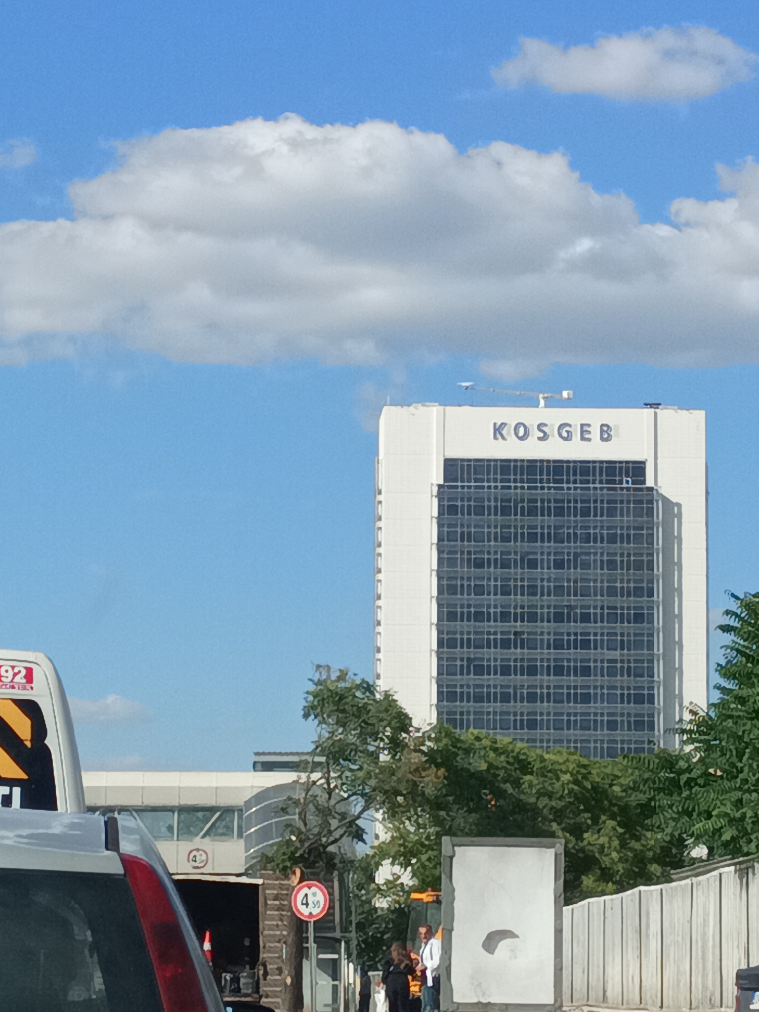 The image shows a tall, modern office building with the name "KOSGEB" prominently displayed at its top. The building has a distinctive glass facade and is surrounded by lush green trees. In the foreground, a vehicle is partially visible, suggesting a street-level perspective. Further in the background, there are other structures, including what appears to be a pedestrian walkway or bridge with signage, and a fence. The sky is bright blue with scattered white clouds, indicating it's daytime, likely a sunny day. Two individuals are standing near a white advertising board or structure in the middle ground, with one person appearing to be a man in a white shirt and another person in dark clothing. The overall scene appears to be an urban or commercial setting in Altındağ, Turkey. There are road signs visible, one with the number "4" and a circle, and another circular red sign with "4 sø" indicating a possible speed limit or restriction. Another sign on the side of the vehicle shows "92" and some other text that is difficult to read. The presence of the KOSGEB building suggests it might be a government or business hub.