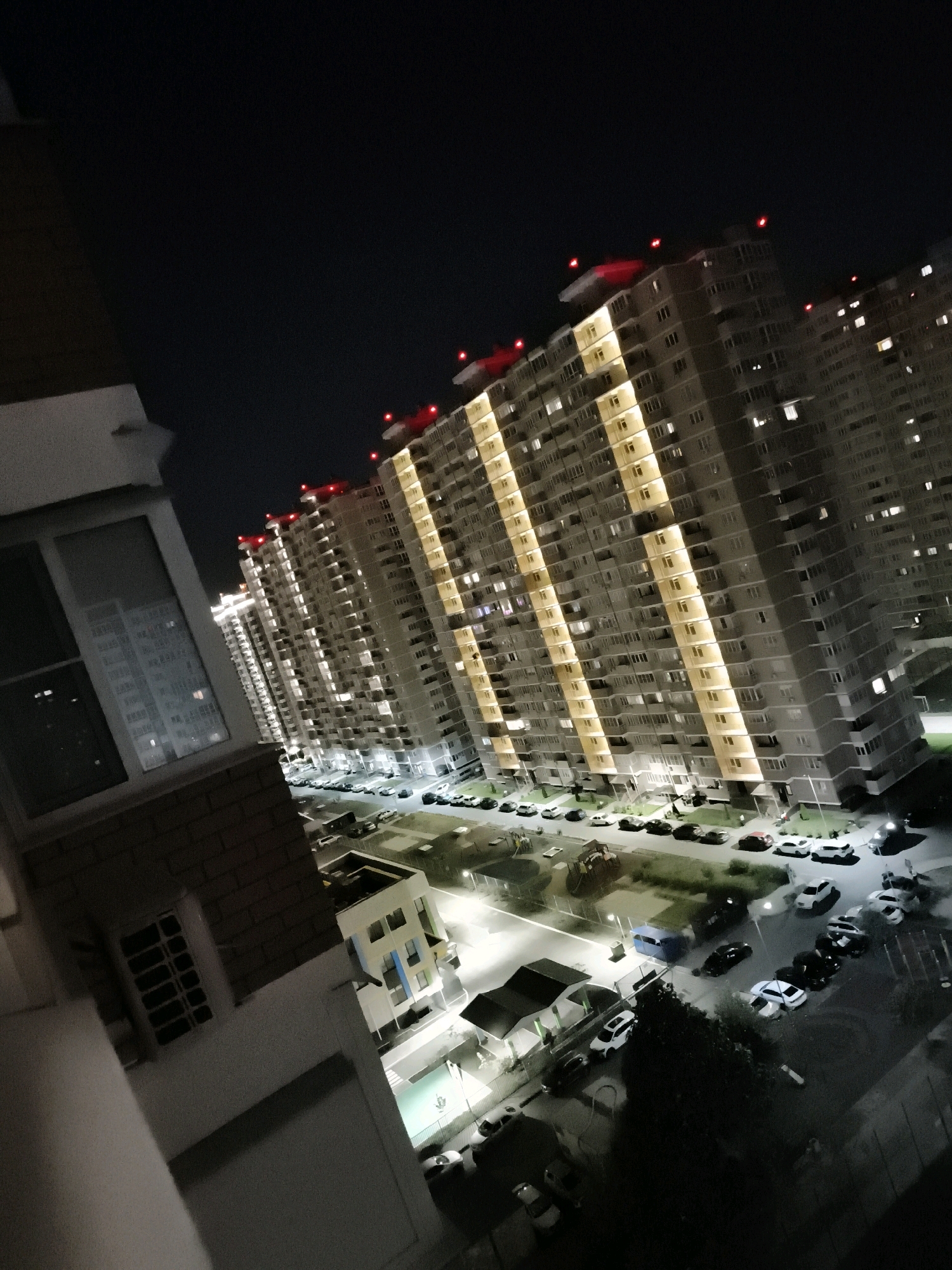 This image shows a nighttime view of a modern residential complex in the city of Krasnodar, Russia. The focal point is a long, multi-story apartment building with a distinctive facade featuring vertical yellow lighting strips that illuminate the windows. Red lights are visible on the rooftop of the building, likely for aviation safety. Below the building, a street is lined with parked cars, and a playground with colorful equipment is visible in the lower right quadrant of the image. Another smaller, modern building with colorful accents is situated to the left, also illuminated at night. The overall scene suggests a well-developed urban environment with contemporary architecture and amenities.