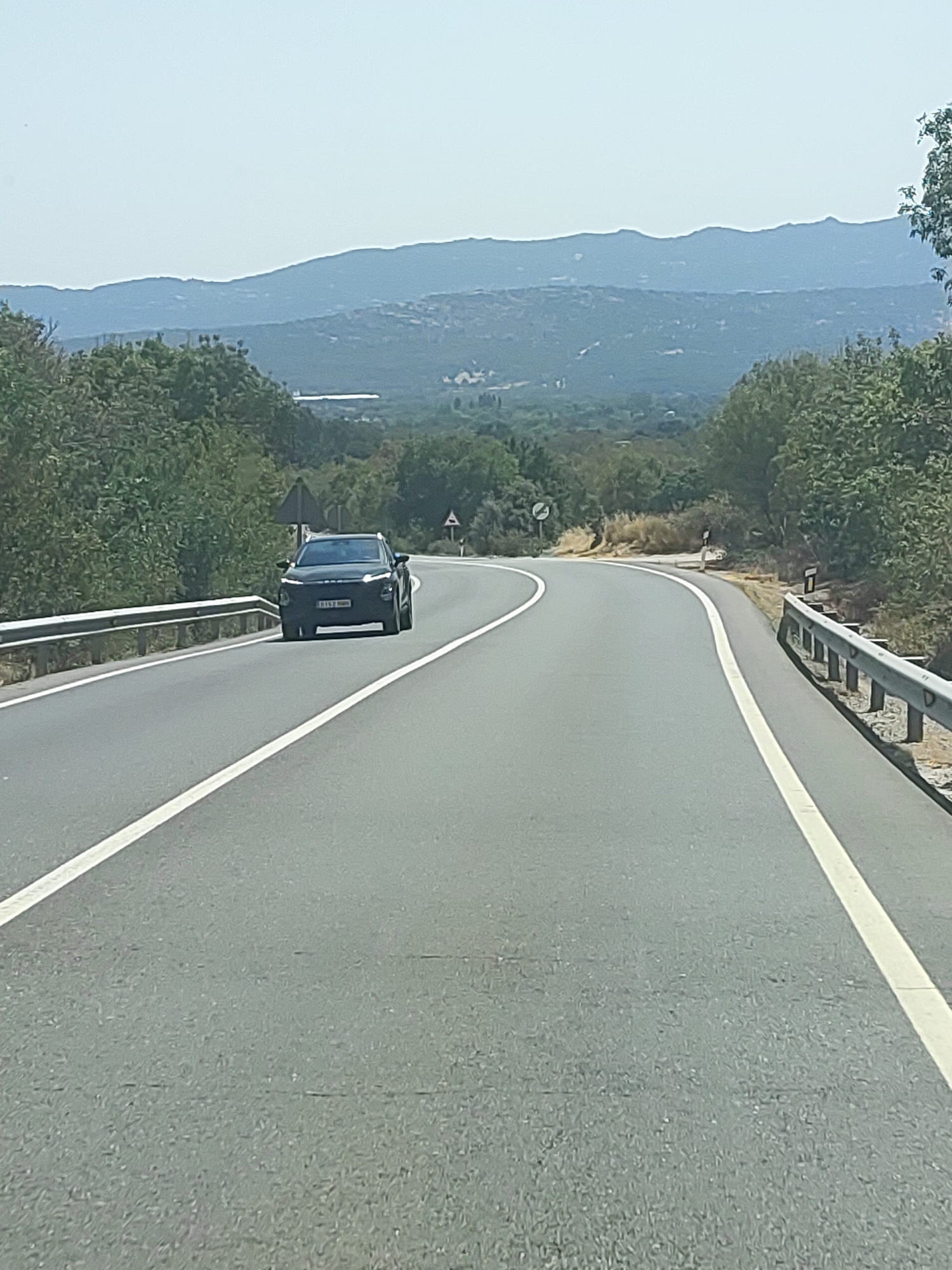 The image depicts a two-lane asphalt road curving to the right, bordered by a metal guardrail on the right and a similar guardrail on the left. Lush green trees and vegetation line both sides of the road, with a hazy, mountainous landscape visible in the background. The sky is a pale blue. A dark-colored SUV is driving on the road, approaching the viewer. The license plate is partially obscured but appears to read "B157 NBB". The scene is captured from a low-angle perspective, suggesting it was taken from a moving vehicle. The environment is consistent with a rural or semi-rural setting. No specific landmarks identifying Becerril de la Sierra are present in the image.