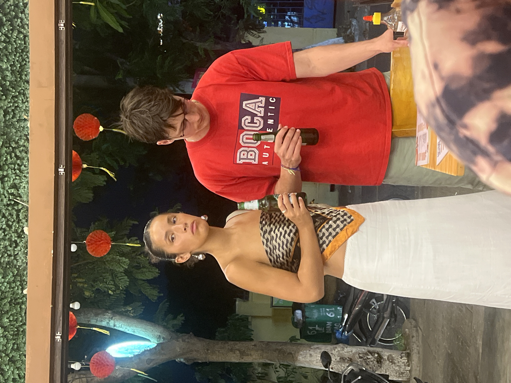 This media file shows two people in what appears to be an outdoor, nighttime setting, likely a street or a patio in Đà Nẵng, Vietnam. The lighting suggests it's after dark, with artificial lights illuminating the scene and creating shadows.
In the foreground, a young woman is shown from the chest up. She is wearing a patterned scarf that is draped around her shoulders and chest, and a white skirt. Her hair is pulled back, and she is wearing large, statement earrings. Her expression is direct and seems serious or thoughtful. She is holding a glass bottle.
Behind her and to the side, a young man is visible. He is wearing glasses and a red t-shirt with the word "BOCA AUTHENTIC" printed on it. He is holding a green bottle. His gaze is directed downwards.
The background shows a lush green, textured wall on the left, possibly a hedge or artificial covering. Further back, there are trees and foliage. Red, circular lanterns are hanging from strings, adding a festive touch to the scene. The presence of motorcycles in the lower part of the frame suggests an urban environment. The overall atmosphere is casual and social.