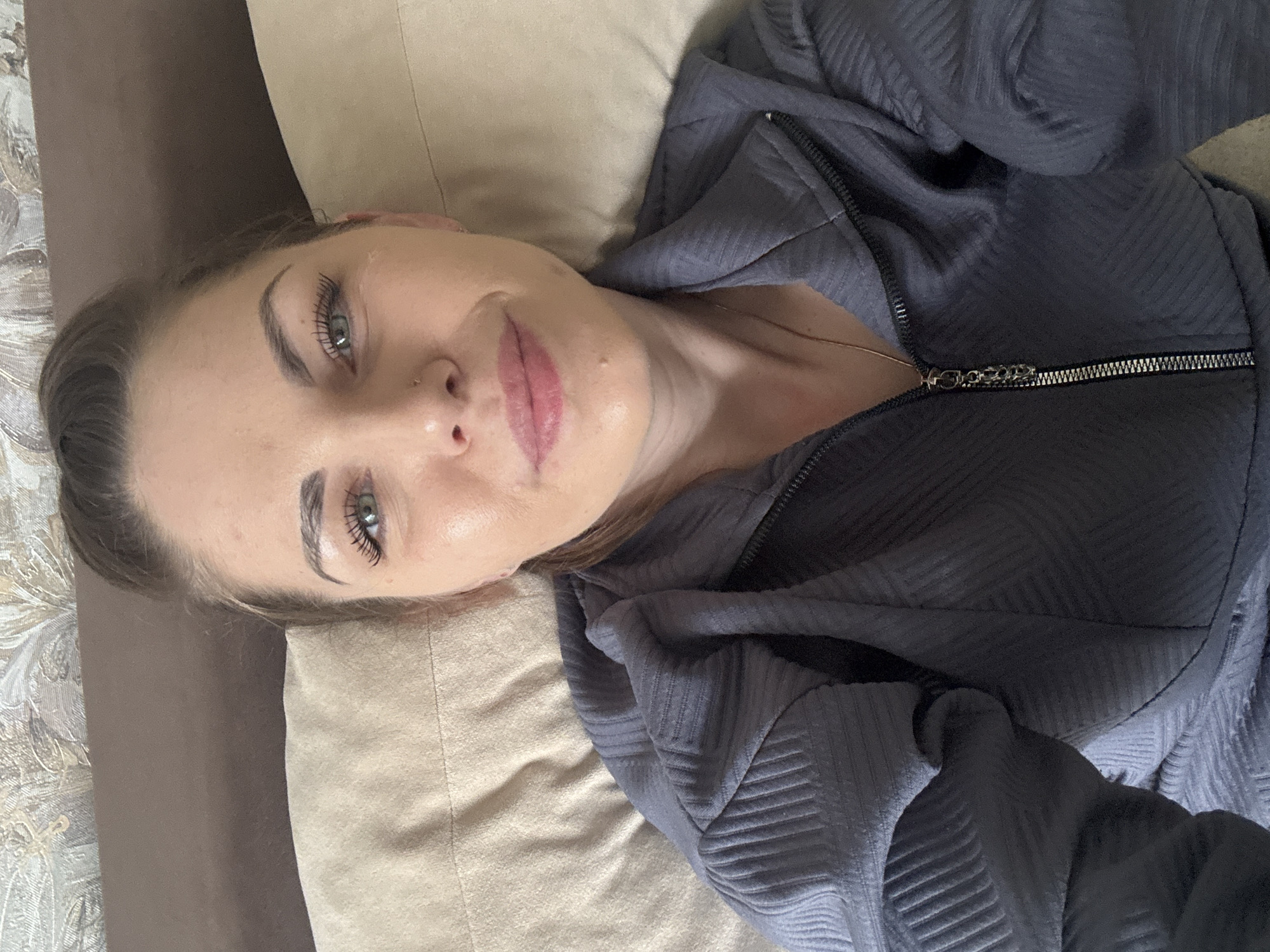 A woman lies on a tan-colored pillow against a floral patterned backdrop, wearing a zippered grey top. She has light skin, green eyes, and a nose piercing.