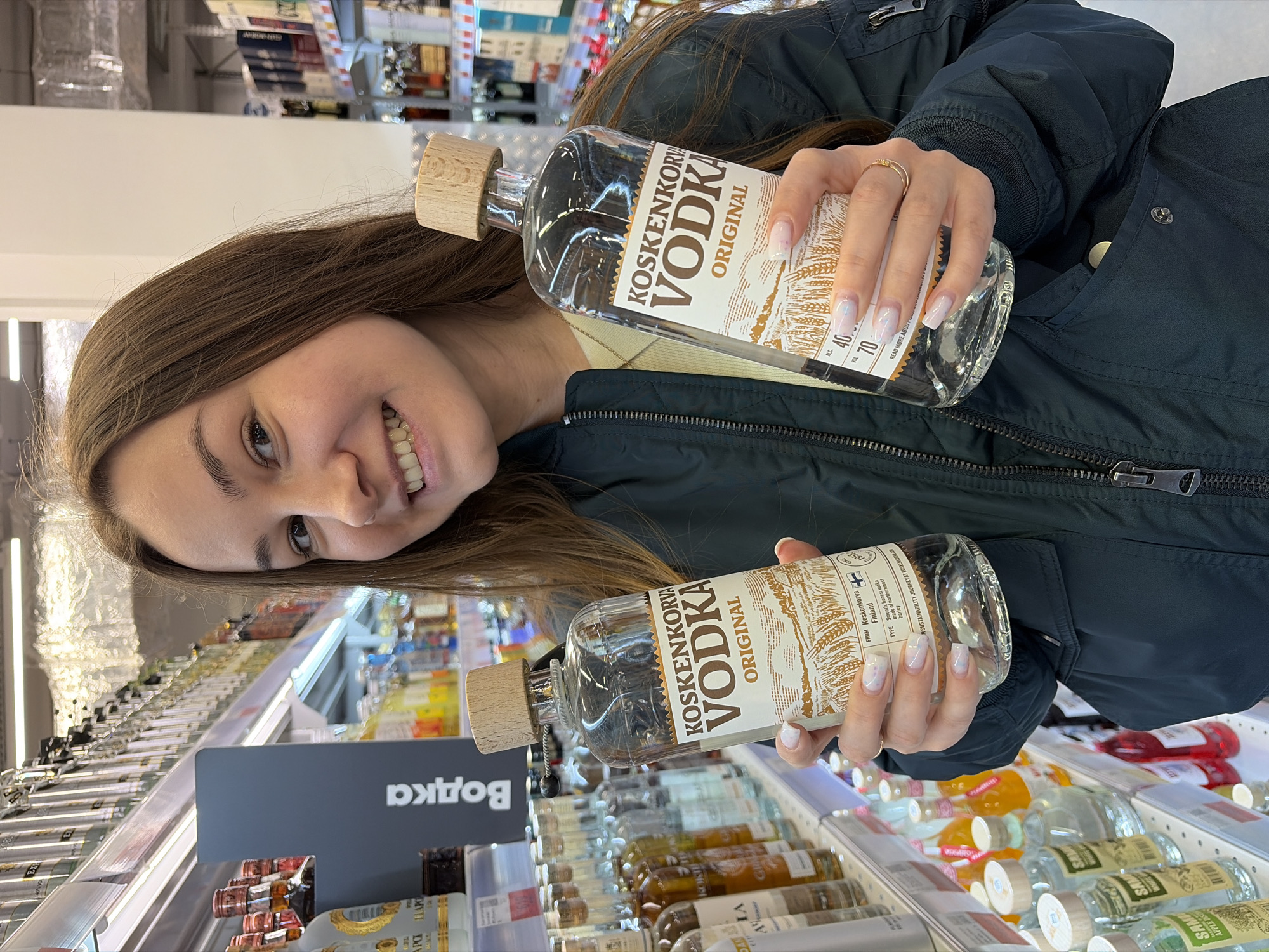 A young woman with light brown hair and fair skin stands in a store aisle, smiling at the camera. She holds two bottles of Koskenkorva Vodka Original, one in each hand. She wears a dark-colored jacket. The store shelves behind her are filled with various alcoholic beverages. A sign with Cyrillic text is visible to the left of the woman.