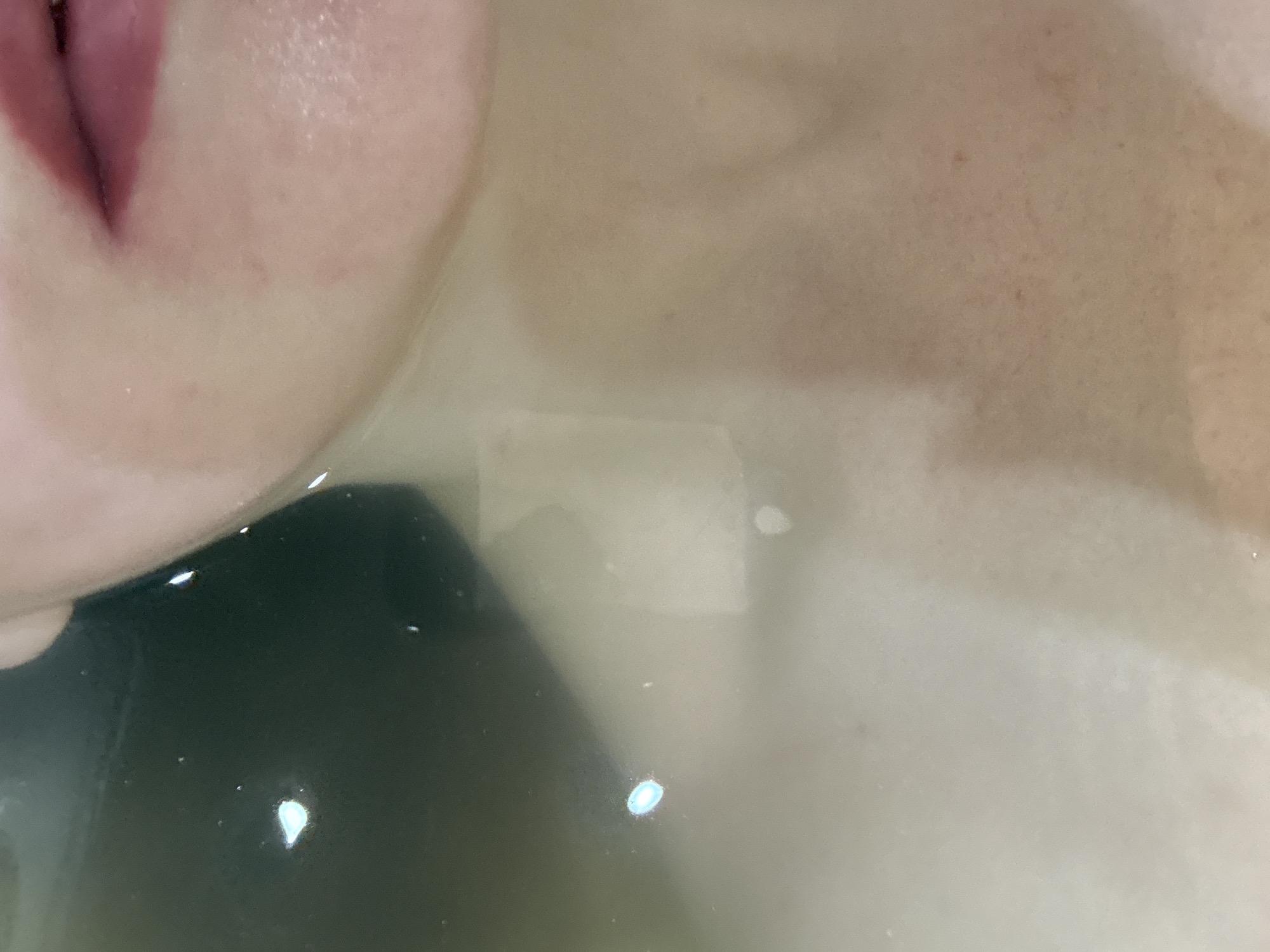 The image shows a close-up of a person submerged in water, with their mouth and part of their face visible above the waterline. The water is somewhat murky, obscuring details of what lies beneath the surface. A dark object, possibly a phone or other electronic device, is submerged in the water. The environment seems to be a bathtub or similar bathing vessel. Location in Chaikovsky, Russia is not evident.