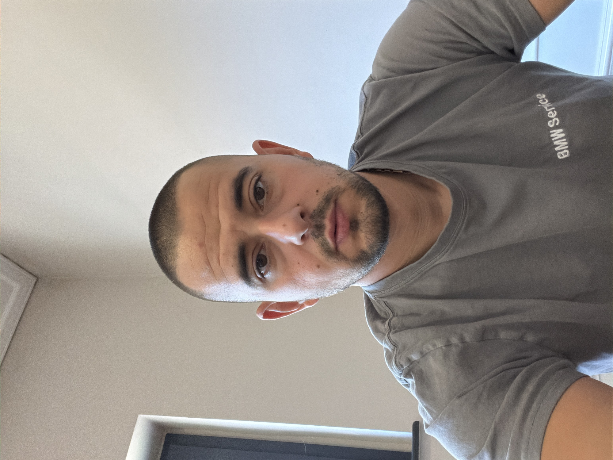 A man with a shaved head and a short beard is looking directly at the camera. He is wearing a grey t-shirt with "BMW Service" embroidered on the left side of his chest. The background is a plain, light-colored wall with some architectural molding at the top. To the left of the man, a dark window frame is visible. The lighting suggests an indoor setting, possibly an office or a workshop. There is no specific information about the location in Burgas, Bulgaria.