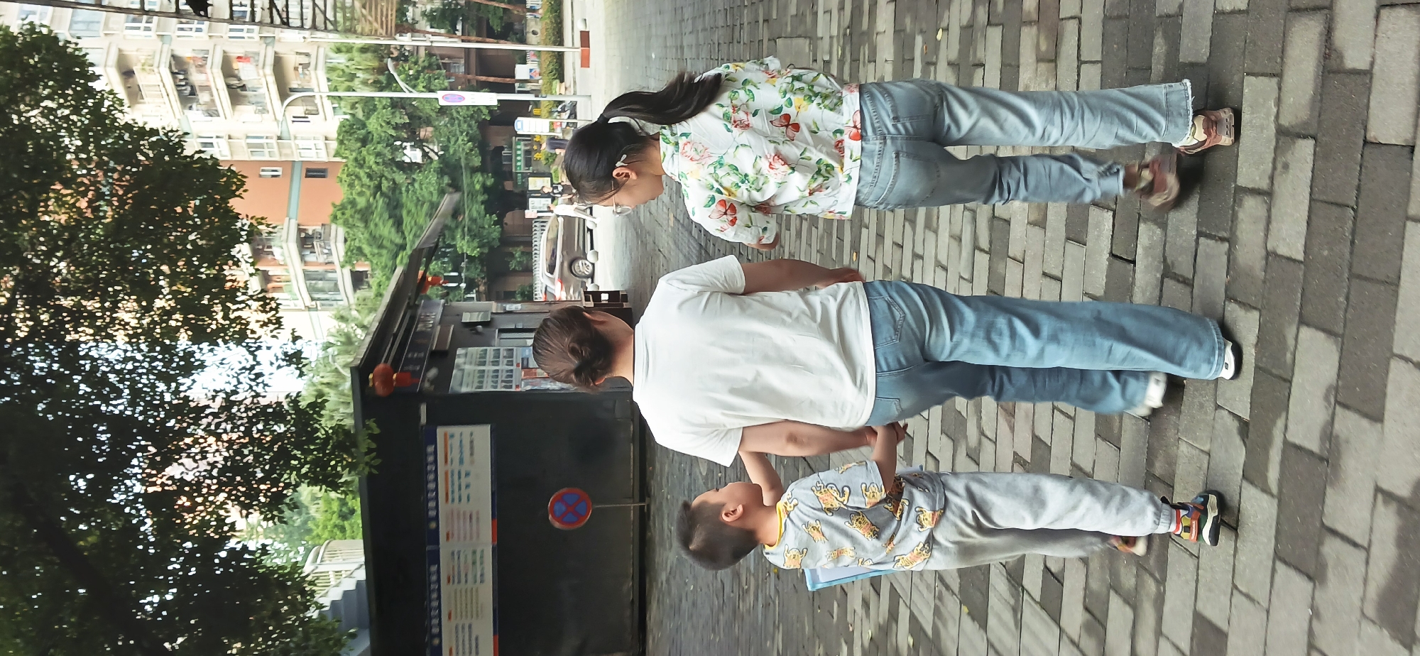 Three people, two adults and a child, stand on a brick walkway in an urban environment. The older woman, nearest the camera, wears a white t-shirt and jeans, with her hair pulled back. The child is wearing a grey tiger print shirt and sweatpants, standing beside the woman. The other adult, a young woman, wears a floral top and jeans, with long dark hair in a ponytail. A grey car is parked in the street. The location appears to be in Jiulongpo District, China, based on signage visible in the background.