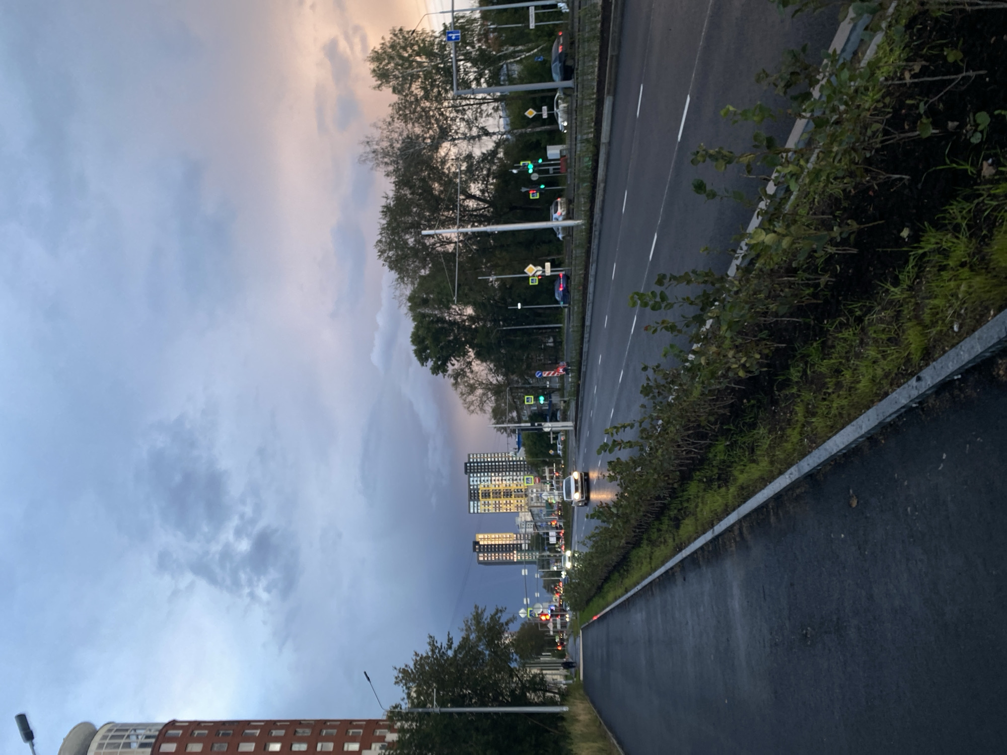 The image depicts a street scene in Perm, Russia, likely taken during twilight. In the foreground, a wet asphalt pedestrian walkway runs alongside a busy road. A low hedge of green bushes separates the walkway from the road, which has multiple lanes and white lane markings. Several cars are visible on the road, some with their headlights on. Traffic lights and street lamps are also present.
In the mid-ground, a line of deciduous trees stands behind the road, with some appearing bare of leaves. Beyond the trees, two modern high-rise buildings dominate the skyline. The building on the left is dark with many windows, while the building on the right is lighter, with yellow accents and illuminated windows.
The sky above is a dramatic mix of dark, stormy clouds on the left, transitioning to lighter, pastel hues of pink and orange on the right, suggesting the setting sun. The overall atmosphere is one of a city transitioning from day to evening, with a hint of impending weather.
