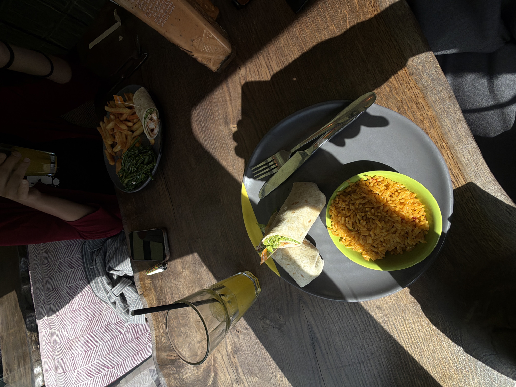 This image shows a wooden table in direct sunlight with two plates of food and two drinks. One plate has a wrap cut in half, a bowl of rice, and cutlery. The other plate has french fries, a wrap, and leafy greens. One of the drinks is in a tall clear glass with a black straw, and the other is in a short glass. A person is sitting at the table, wearing a patterned shirt and a red shirt. There is also a cell phone and a lighter on the table. A bottle is visible at the top-left of the frame, with the label partially visible. The location is not discernible based on the objects in the image.
