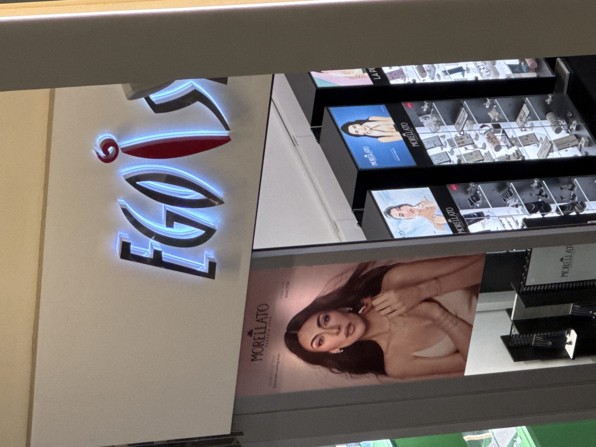 A vertically-oriented eye-level shot depicts an interior scene, likely a store, in Yerevan, Armenia. In the foreground is a storefront display with the name "MORELLATO" appearing vertically on a sign alongside a large portrait of a woman with dark hair wearing jewelry. Above this, there is an illuminated sign of the brand name "FOLLIE" in large, stylized letters, with the letters lit by a cool blue light. Display cases with additional "MORELLATO" branding contain jewelry and watches. The overall lighting in the store is bright and modern.