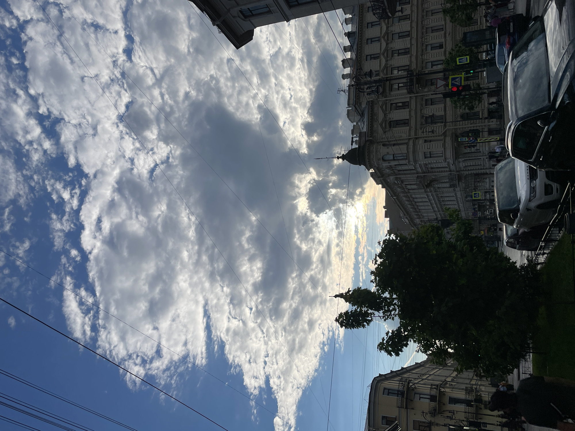 The image captures a vibrant street scene in Saint Petersburg, Russia, under a dynamic sky. The sky dominates the upper portion of the frame, filled with large, fluffy white and grey clouds, interspersed with patches of clear blue sky. Sunlight filters through and behind the clouds, creating bright highlights and casting softer light on parts of the scene. Multiple thin overhead power or tram lines crisscross the sky.
On the right side of the image, ornate, multi-story historical buildings line what appears to be a busy city street. One prominent building features an elegant domed cupola. Several cars, including a white SUV with "RANGE ROVER" visible on its rear, are parked along the curb or moving on the street. Lush green trees with dense foliage are scattered along the sidewalk, providing shade.
The lighting suggests late afternoon or early evening, with the sun low in the sky, creating a mix of bright and shadowed areas. A red traffic light is visible further down the street, and pedestrian crossing signs are also discernible. A few blurry figures of people can be seen on the sidewalk. The architecture points to a classic European urban setting, consistent with the specified location of Saint Petersburg.