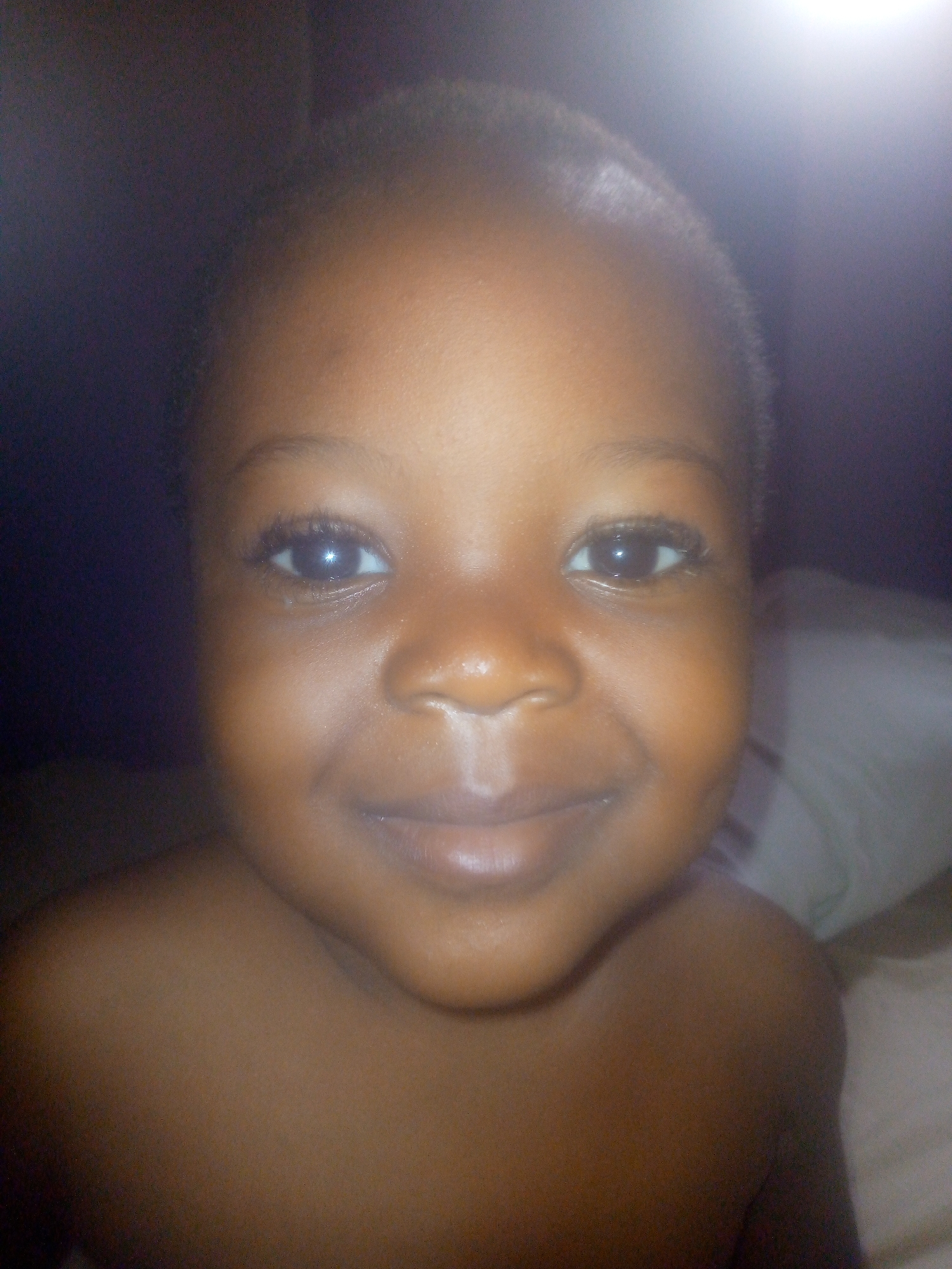 A close-up shot features a child with dark skin and short, dark hair. The child is facing the camera with a slight smile. The background is dark and indistinct, with what appears to be a pillow partially visible on the right side. The location is not specified.
