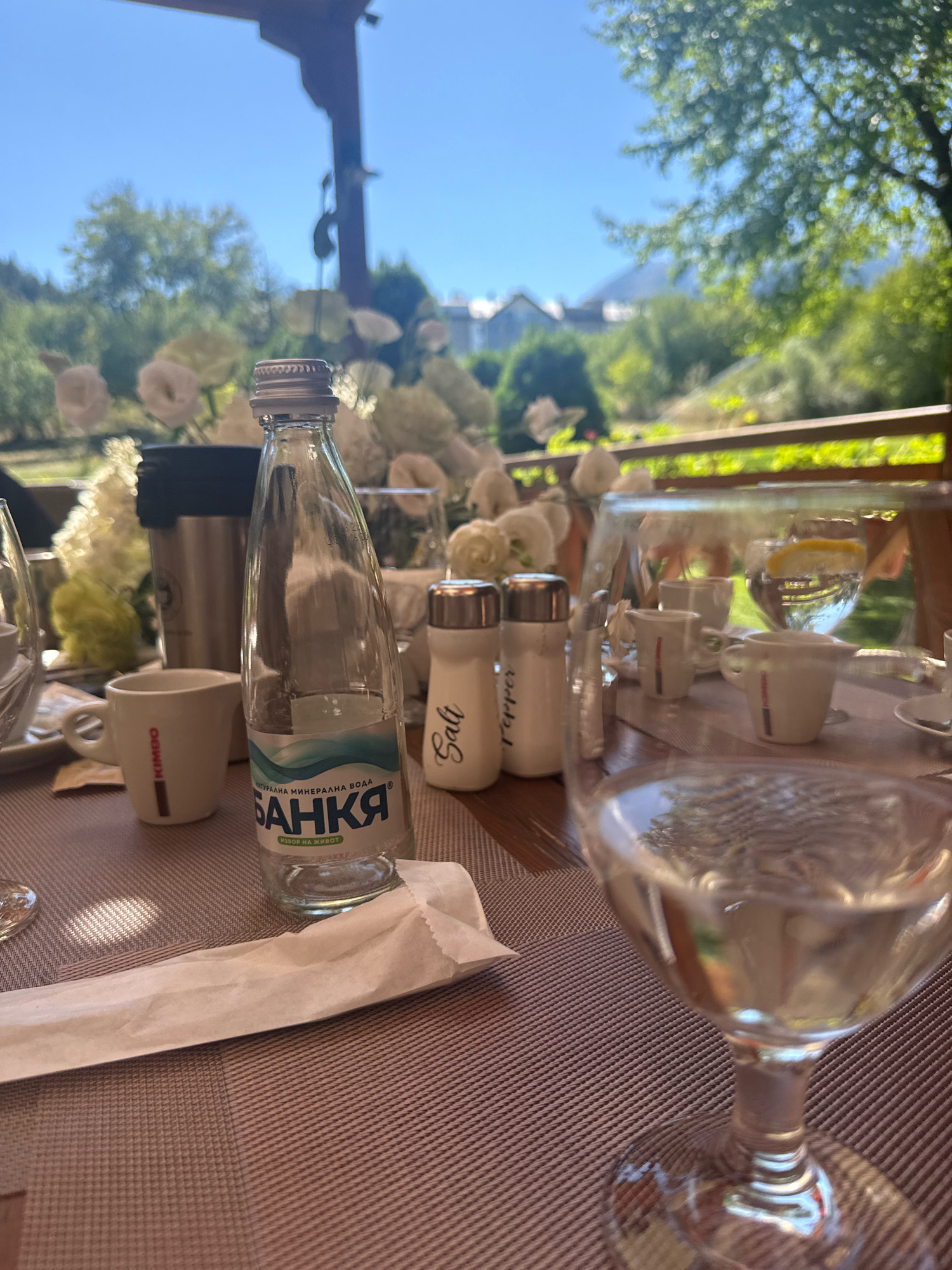 This is a close-up, eye-level shot of a table set for a meal, likely outdoors, with a backdrop of trees and buildings in the distance. The table is covered with a brown textured placemat. In the foreground, a wine glass filled with a clear liquid is prominently featured. To the left of the glass, a bottle of "BANKA" mineral water stands, with a portion of its label visible, displaying text in Cyrillic. Next to the water bottle are salt and pepper shakers. Further to the left, a small white cup, possibly for espresso, with "KIMBO" branding, is visible. In the background, a blurred arrangement of white flowers adds a decorative element. Beyond the table setting, a wooden railing suggests an outdoor seating area, possibly a terrace or balcony, with lush green trees and buildings under a clear blue sky. The overall impression is a serene outdoor dining setting in Bansko, Bulgaria, with elements of a meal and refreshments.