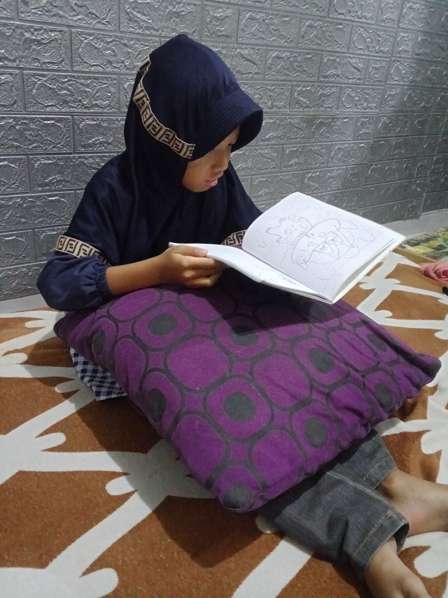 The image depicts a young person, likely a child, wearing a dark blue hijab with a beige logo trim and a dark blue long-sleeved shirt. They are seated on a bed or floor covered with a brown and white patterned blanket, leaning against a large purple pillow with a black circular pattern. The person is holding an open book with a drawing on its pages and appears to be engaged in reading or drawing. The background shows a textured grey brick wall. The scene is indoors, likely in a residential setting in Kemulan, Indonesia.