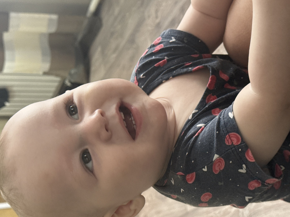 The image displays a close-up, eye-level view of an infant with light skin, dark eyes, and a smooth scalp, positioned indoors. The infant's head is tilted slightly upwards and to the left, with eyes directed towards the upper right. An open-mouthed smile reveals visible lower teeth. A faint, small mark is present on the infant's forehead above the right eyebrow. The infant is wearing a dark-colored, short-sleeved garment patterned with red circular motifs and smaller white heart shapes. Portions of the infant's arms and knees are visible in the lower frame. The background consists of light brown wooden plank flooring in the immediate foreground, transitioning to a blurred, light-toned area with vertical stripes, possibly a wall or window covering. No visual information within the image identifies the location as the city of Кипчак, Russia.