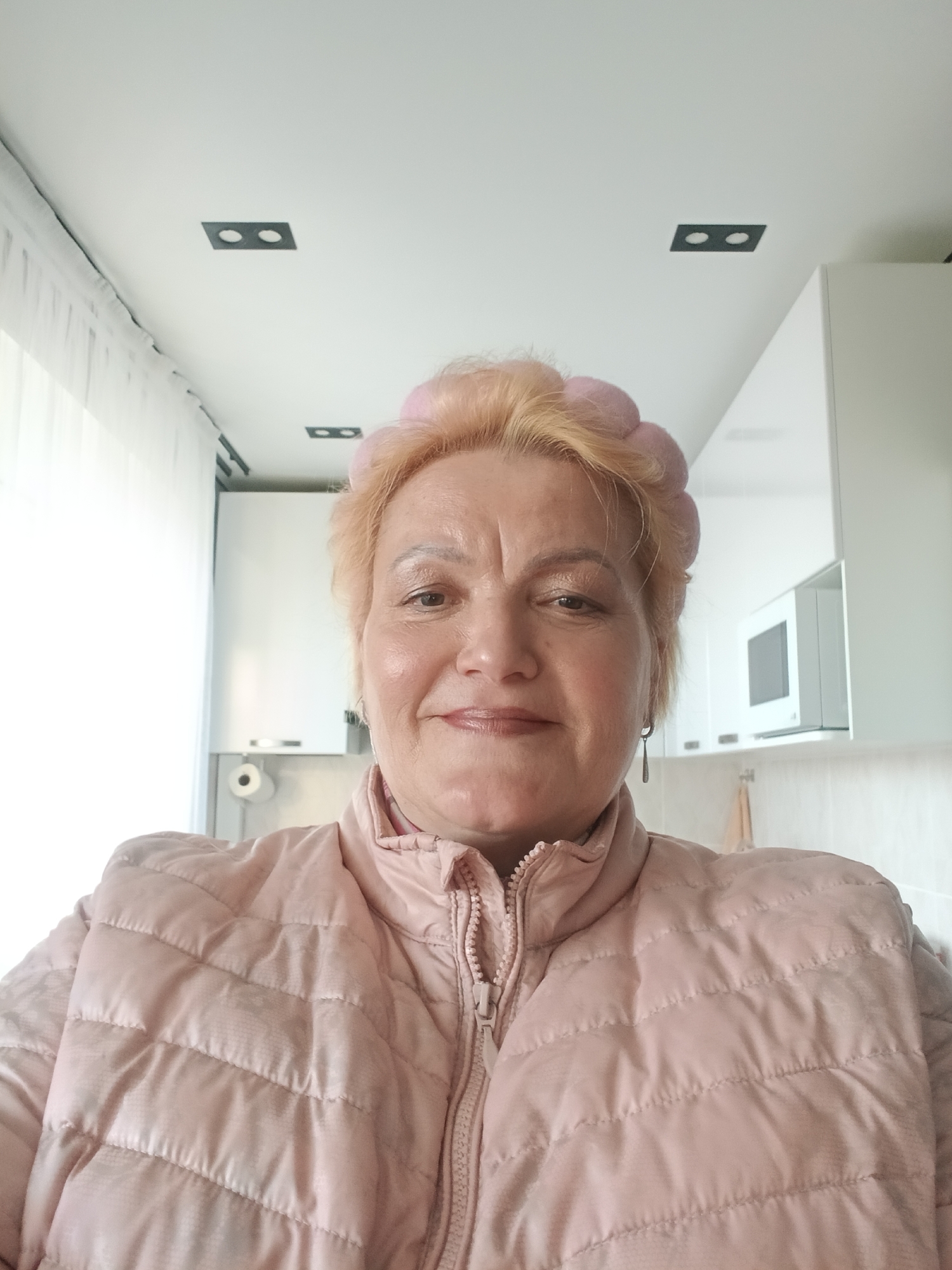 A woman with short blonde hair and a pink padded jacket is indoors. She is wearing a pink headwrap and earrings. Behind her is a white kitchen with cabinets, a microwave, and what appears to be a paper towel dispenser. The ceiling is white with two rectangular light fixtures with two lights in each. A window with white curtains is visible on the left. The location is potentially in Murom, Russia.