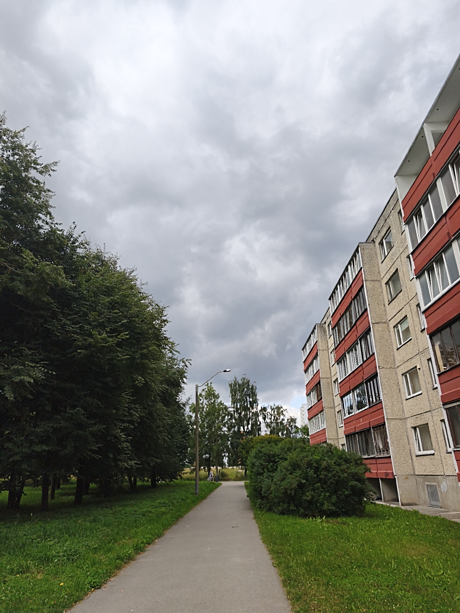 This is a full, outdoor, medium-shot, eye-level, slightly tilted photograph taken on a cloudy day. The scene depicts a pathway between a row of trees and a modern, multi-story apartment building. The building, on the right, is concrete with red and white balconies and windows. The trees, on the left, are tall and leafy. The pathway is paved and leads into the distance. There are no people visible. The sky is overcast with heavy gray clouds. The overall environment suggests a residential urban area in Tallinn, Estonia.