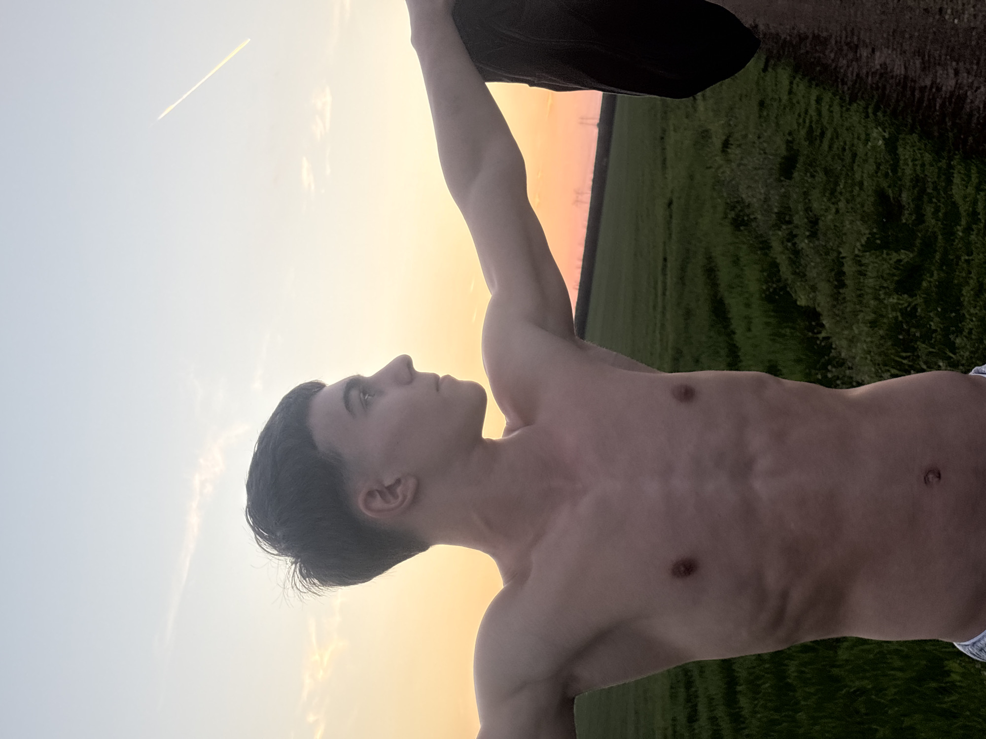 A shirtless, fair-skinned young man stands in a field under a sunset sky. He is looking upwards and to the left. His left arm is raised, and he appears to be holding an object above his head, possibly a piece of clothing. The field is green, and there are utility poles visible on the horizon. A contrail is present in the sky. The location is near the city of Lehchevo, Bulgaria.