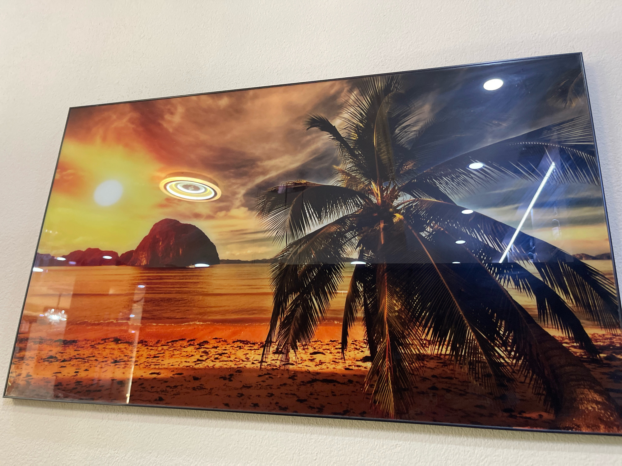 The image is a photograph of a framed piece of art hanging on a textured white wall. The artwork depicts a tropical beach scene at sunset, with a prominent palm tree in the foreground. The sky is a vibrant orange and yellow, with dramatic clouds. In the distance, a large rock formation rises from the ocean, and the sun is setting. The beach is sandy, with some debris or seaweed scattered on it. The artwork is glossy and reflects light, with visible glare from overhead lights. The framing is thin and black. The location of the artwork in the city of Mirushë, Kosovo, is not discernible from the image itself.