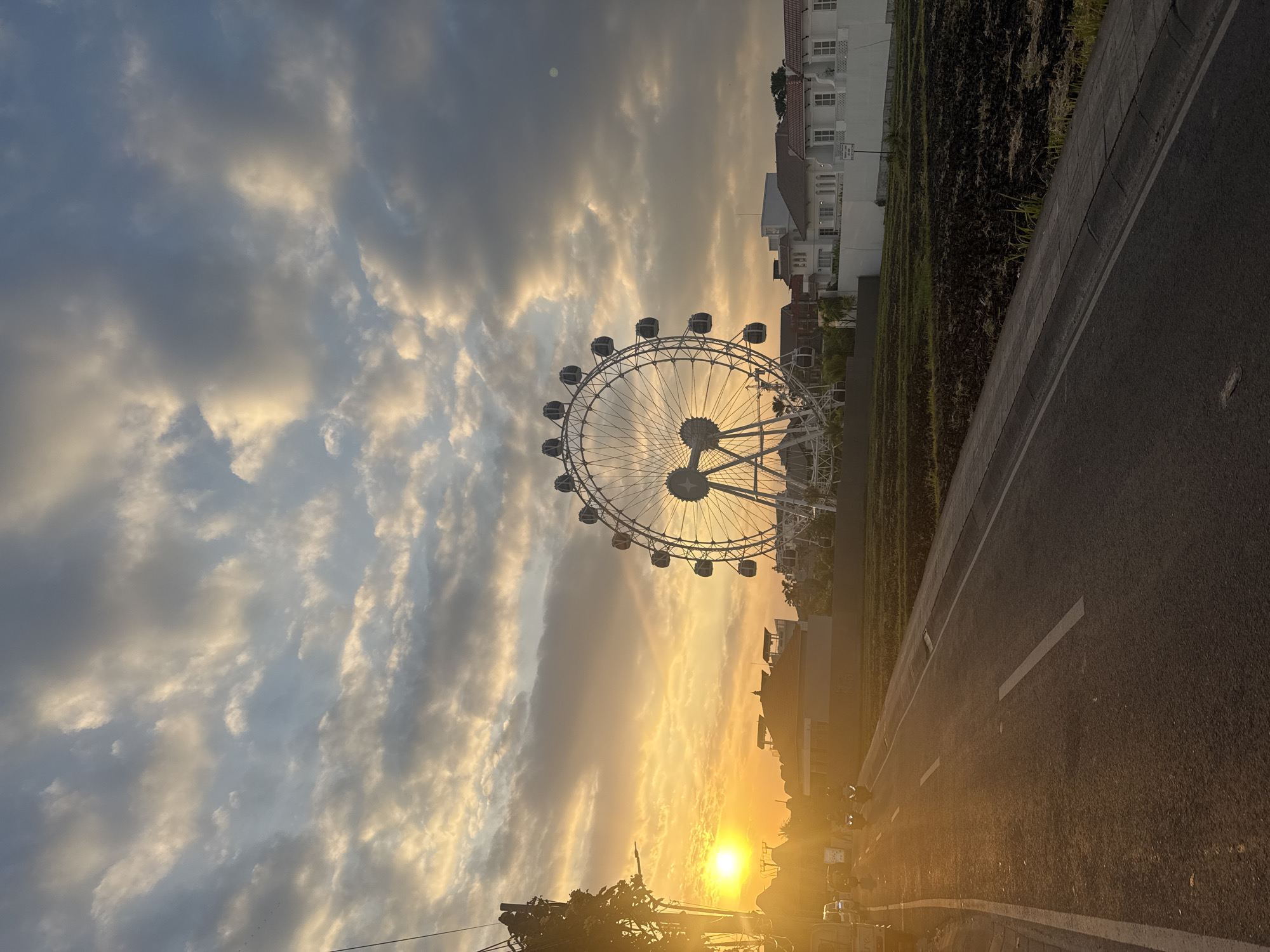 This image captures a sunset scene in North Kuta, Indonesia. A large Ferris wheel is the dominant structure in the mid-ground, silhouetted against a dramatic sky filled with clouds tinged with the orange and yellow hues of the setting sun. The sun itself is a bright orb on the left side of the frame, casting a warm glow. In the foreground, a paved road with white lane markings runs diagonally from the bottom left to the bottom right. There are a few vehicles, including motorcycles and cars, visible on the left side of the road. Beyond the road, there are some buildings, including a row of white houses with red-tiled roofs on the right, and what appears to be cultivated land or a field in the mid-ground. Trees and power lines are visible on the far left of the image, further framing the scene. The overall impression is one of a tranquil evening in a developed area with natural and man-made elements.