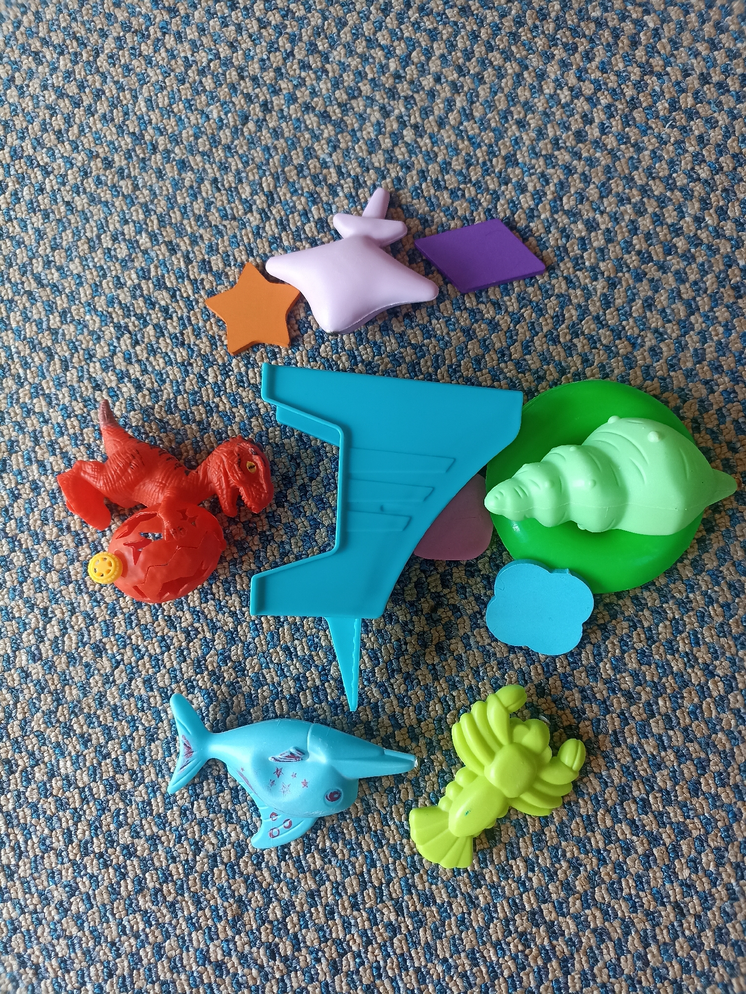 The image displays a collection of colorful children's toys scattered on a blue and beige patterned carpet. The toys include a red dinosaur figure, an orange star, a purple diamond shape, a lavender star, a light blue dolphin, a light green lobster, a light green shell on a green base, and a blue cloud-shaped cutout. A large, bright blue plastic structure with a curved top and straight sides is also visible. The lighting suggests an indoor setting, possibly a child's playroom or living area. There are no people or any other contextual elements that would indicate a specific location within Mojopanggung, Indonesia.