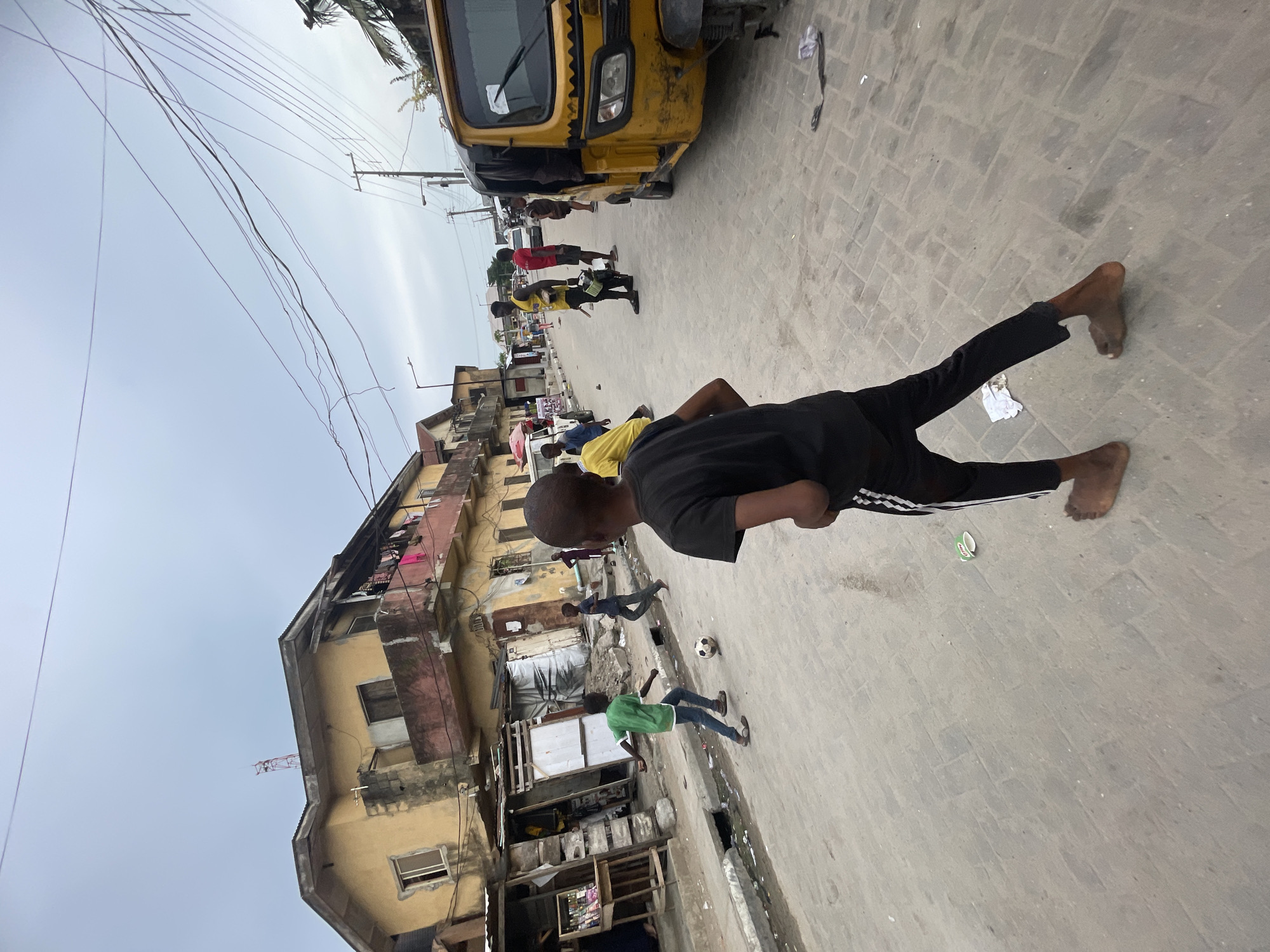 The image, which appears to have been taken in portrait orientation and then rotated, depicts an outdoor street scene in Ado, Nigeria, under an overcast sky. The paved street, featuring rectangular blocks, is the central focus, flanked by multi-story buildings on both sides. In the foreground, a young boy wearing a black t-shirt and black pants with white stripes is seen from behind, standing or walking on the street. Further down the street, several other children are visible, including one in a green t-shirt near a soccer ball, indicating that a game of soccer is likely taking place.
A yellow tricycle (keke napep) is parked or stationary on the right side of the street, with a few adults or older youths standing around it. The buildings along the street show signs of age and use, with laundry hanging from upper floors and exposed electrical wiring. A dense network of utility lines crisscrosses the light grey, overcast sky above. The street surface also shows some scattered litter, such as pieces of paper and a small plastic cup. Many individuals in the scene are barefoot, suggesting a casual and warm environment. No prominent or legible text is visible in the frame.