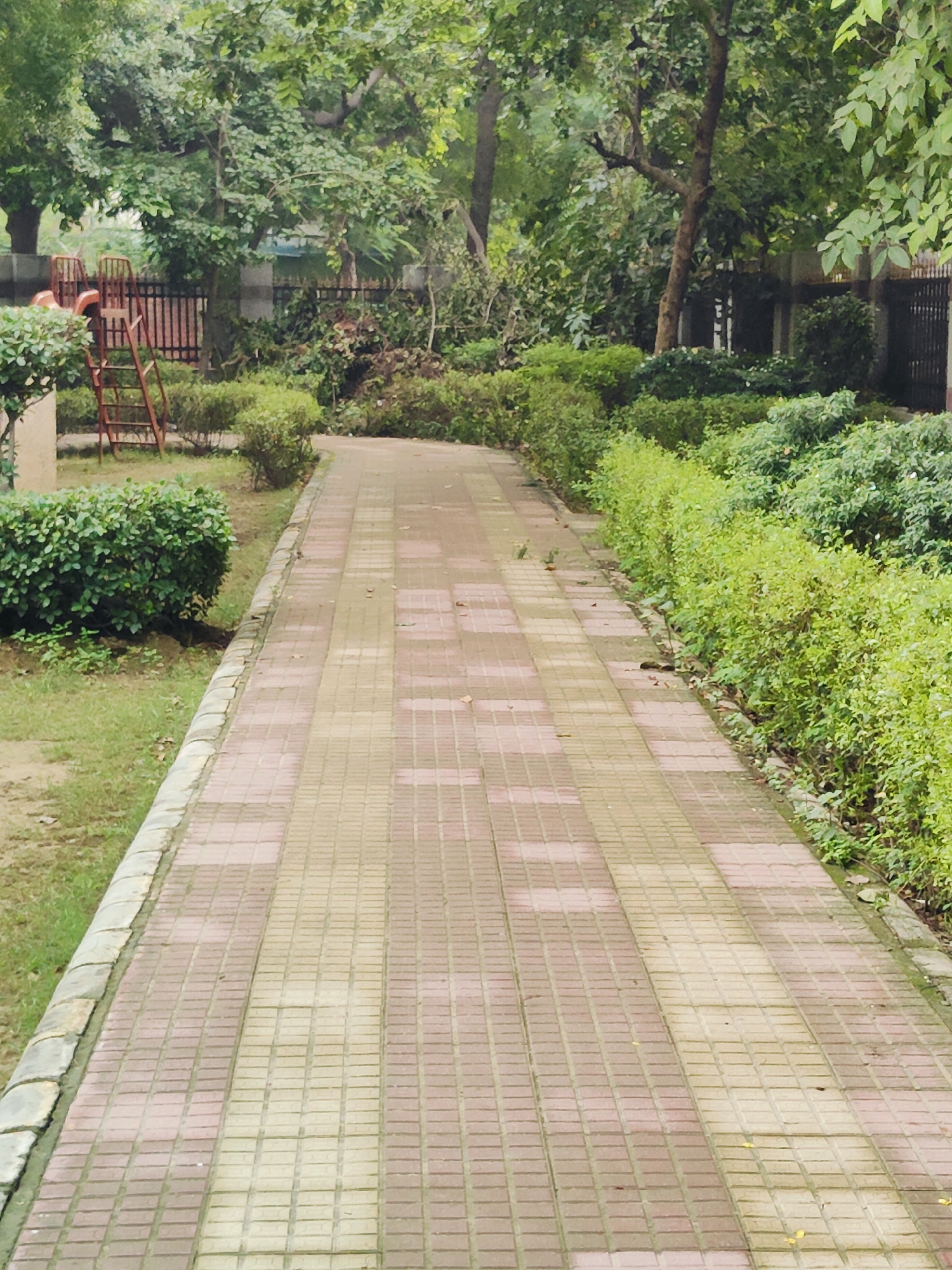 The image shows a paved pathway with a patterned design of red and beige square tiles, curving through a park-like setting. On the left side of the path, there is a grassy area with manicured green bushes. Further back on the left, a red metal slide structure with a ladder is visible behind some foliage. On the right side of the path, there are rows of lush, neatly trimmed green bushes. The background is filled with mature trees and dense green foliage, suggesting a well-maintained urban park. The lighting indicates an overcast day. The scene is likely located in a residential area or public park within Gurgaon, India, given the typical landscaping and playground equipment found in such locations. No people are visible in the image.