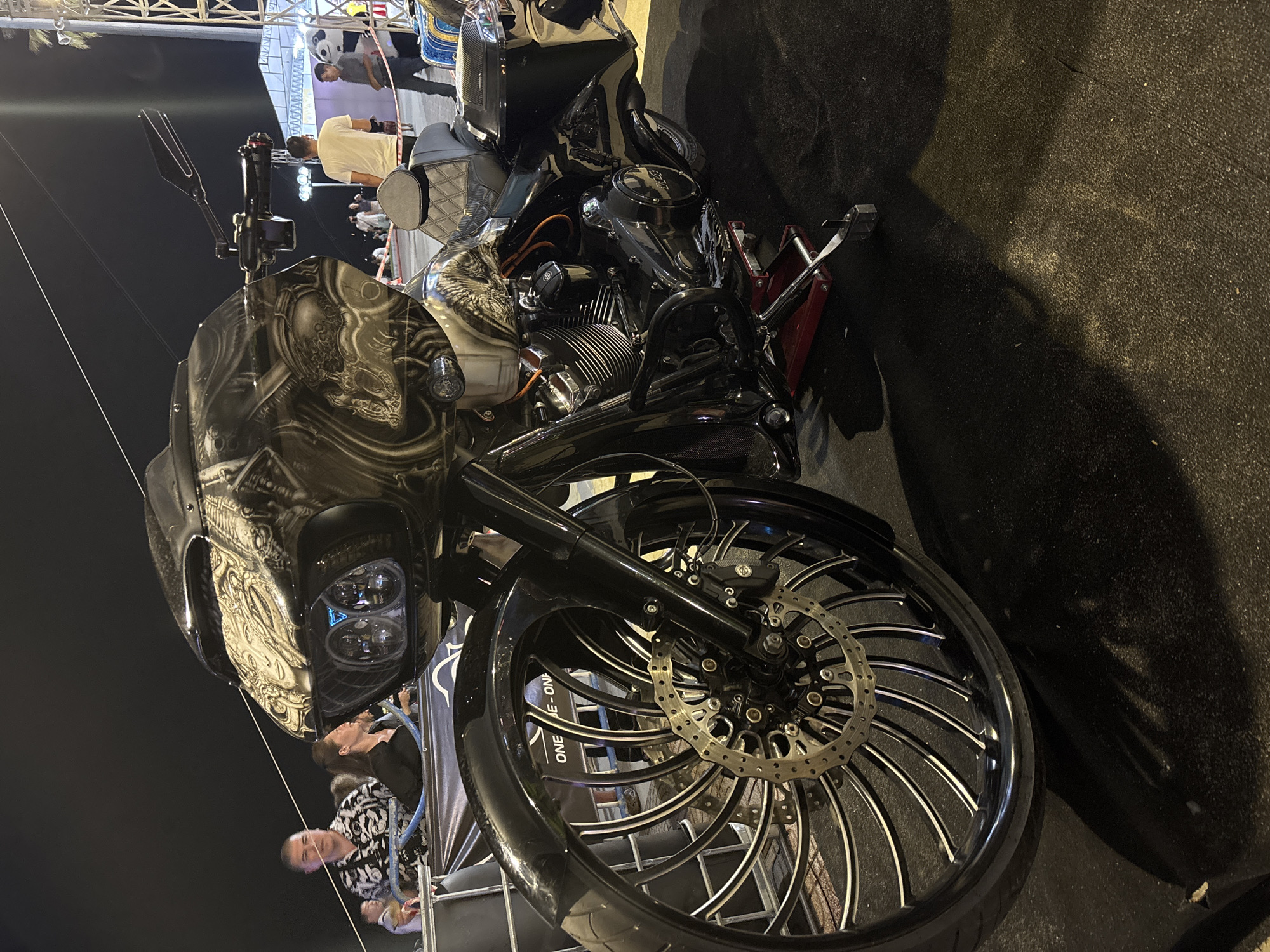 A customized black motorcycle with elaborate airbrush art is displayed. The motorcycle features a large, custom front wheel with a complex spoke design and a dual disc brake system. The front fairing is adorned with skull or skeleton artwork. The motorcycle is positioned on a jack. Several people are standing nearby. The location appears to be a public area, possibly an event or exhibition in Nha Trang Ward, Vietnam.