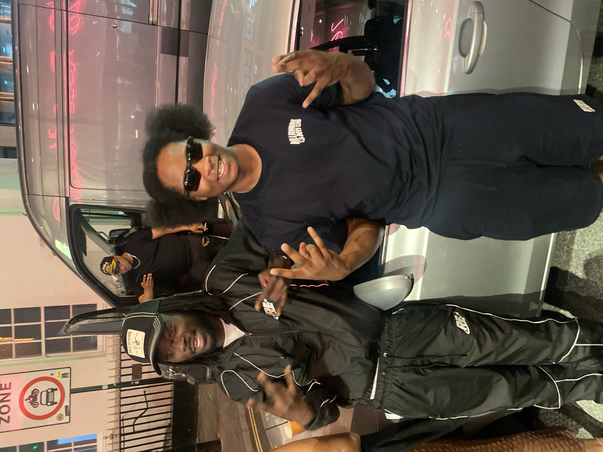 The image shows three individuals posing in front of a silver vehicle. One individual with dark hair, sunglasses, and a dark shirt with "BILLIONAIRE BOYS CLUB" emblazoned on it is throwing up a hand sign. Another individual in a black jacket and cap, with long hair, is also throwing up a hand sign. A third individual can be seen through the open car door. There is a "NO ZONE" sign with an icon of a motorcycle and a car. The setting seems urban with building windows visible in the background. Based on the type of sign and building style, the location is likely in London, United Kingdom.