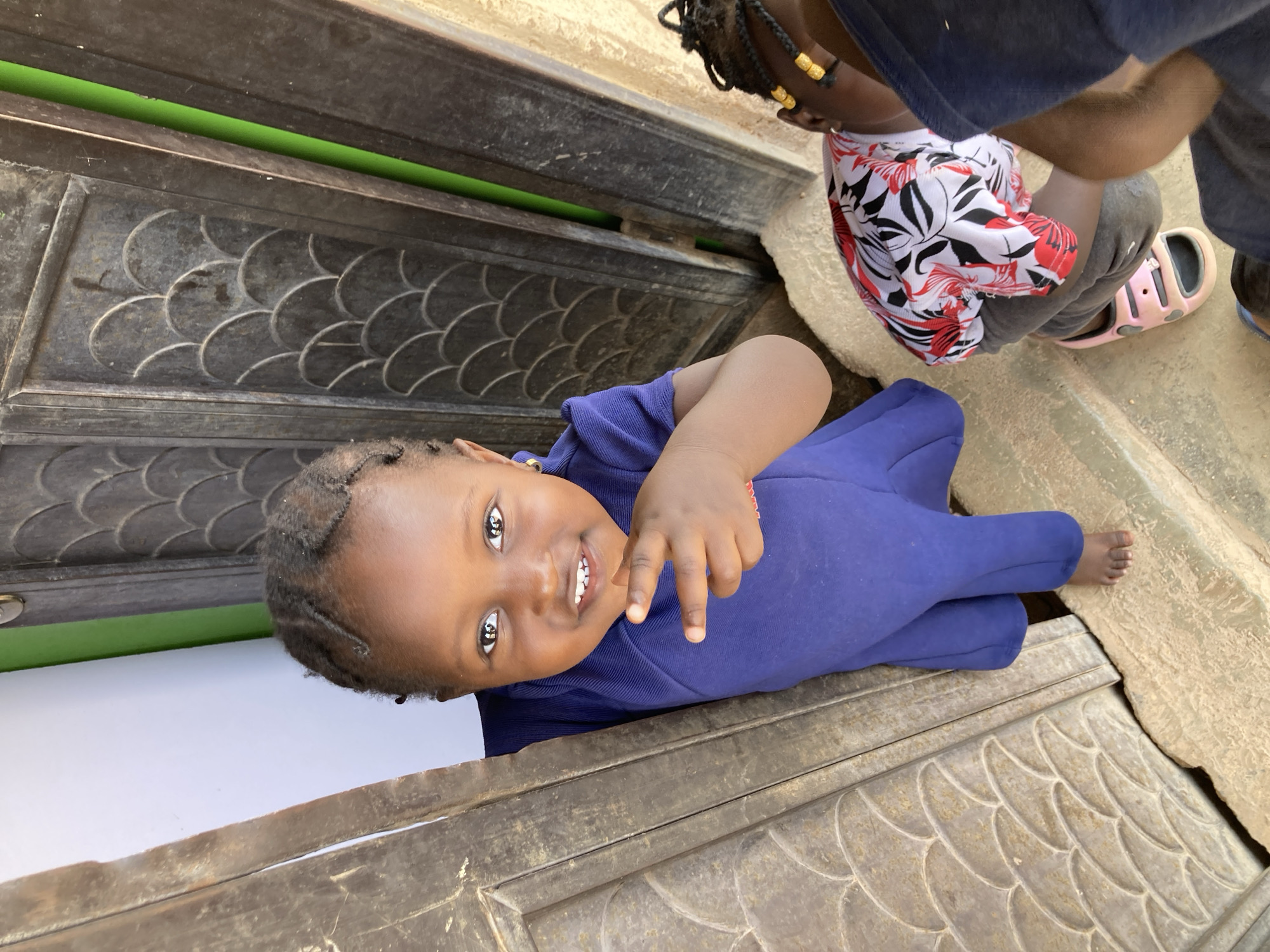 This image captures a lively outdoor scene in Jos, Nigeria, featuring two young children.
The primary subject is a young child with dark skin, positioned in the foreground and looking directly at the camera with a joyful, wide smile that reveals several small teeth. They are wearing a long-sleeved, full-body outfit in a vibrant royal blue color. Their hair is styled in small braids, some of which are adorned with gold beads. The child extends their left hand towards the camera, making a playful gesture, possibly a peace sign. They appear to be leaning or lying on a light-colored, textured ground surface.
Partially visible in the upper right background is another child or person. This individual wears a white top with a distinct red and black floral pattern, paired with grey pants and light pink, iridescent sandals. Their hair also features braids with gold beads. They appear to be bending over or engaged in an activity just outside the frame.
The background is dominated by dark, ornate metalwork, likely part of a gate or door, which features a decorative, repetitive scallop or fish-scale pattern. A narrow strip of bright green is visible through a gap in this metal structure. The ground around the children is a rough, light-colored material, possibly concrete or stone.
The scene is brightly lit by natural daylight, suggesting clear weather conditions. The visible child's smile and gesture convey a sense of happiness and playfulness. There is no visible text in the image.