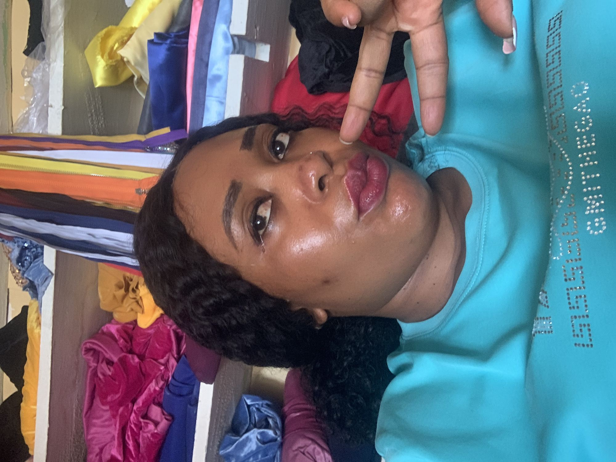 A selfie of a Black woman indoors with medium-length black curly hair, wearing a turquoise shirt with rhinestone lettering. She is making a hand gesture near her face with painted fingernails. Her lips are red and shiny. Behind her is a wall-mounted shelving unit containing fabric, zippers, and sewing supplies. The location is Sabon Tasha, Nigeria.