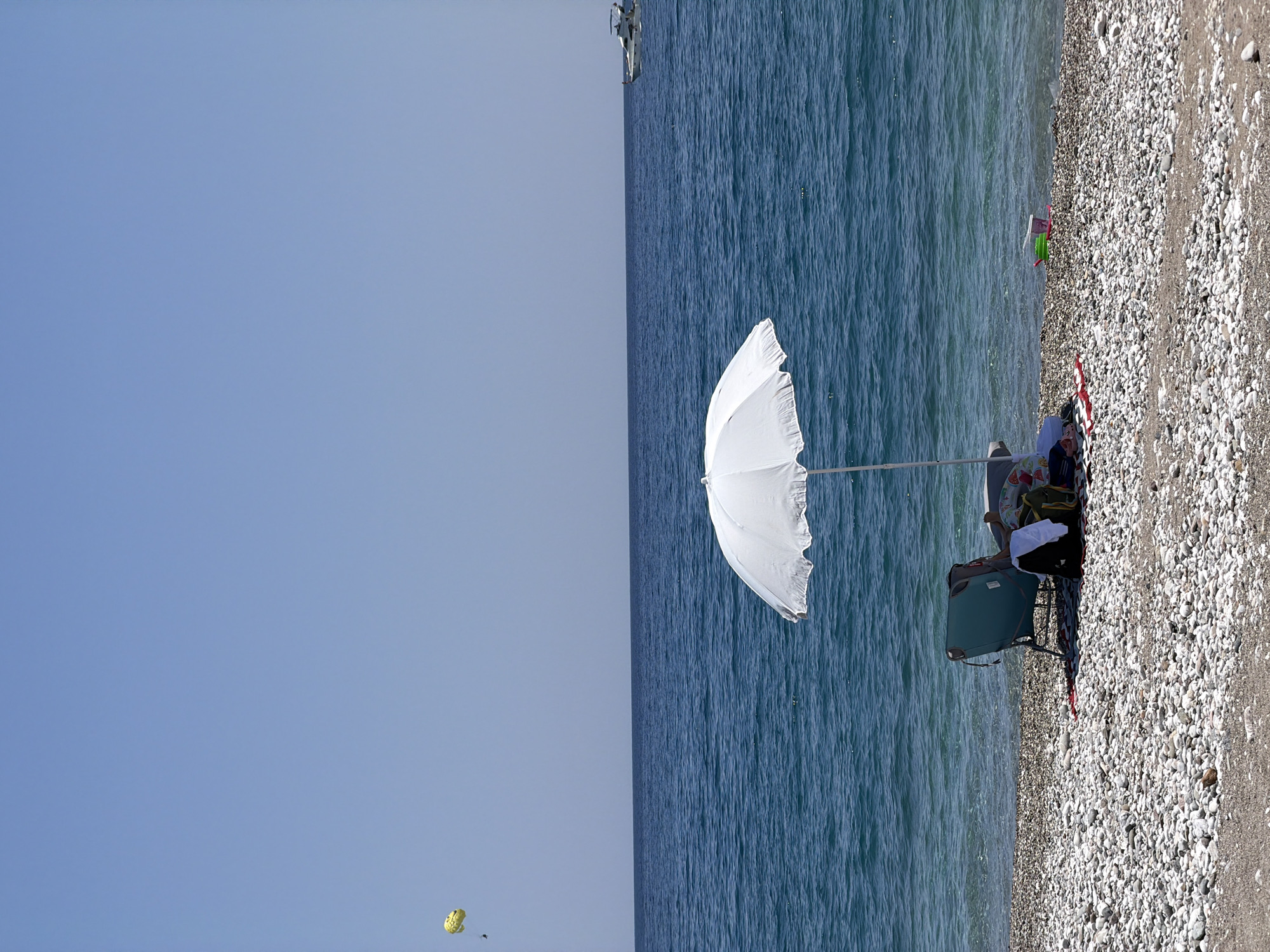 The image captures a serene beach scene in Kemer, Turkey, under a clear blue sky. The foreground is dominated by a pebble beach, leading to the calm, turquoise waters of the Mediterranean Sea. A white beach umbrella provides shade for a teal-colored beach chair, on which a person is seated, partially visible. Next to the chair are a green backpack and a black garment, suggesting personal belongings. A red toy shovel and pail are placed on the sand near the water's edge, indicating children's play. In the distance, a boat cruises across the sea, and further to the left, a yellow parasail with a person attached floats in the sky. The overall ambiance is one of leisure and relaxation on a sunny day.